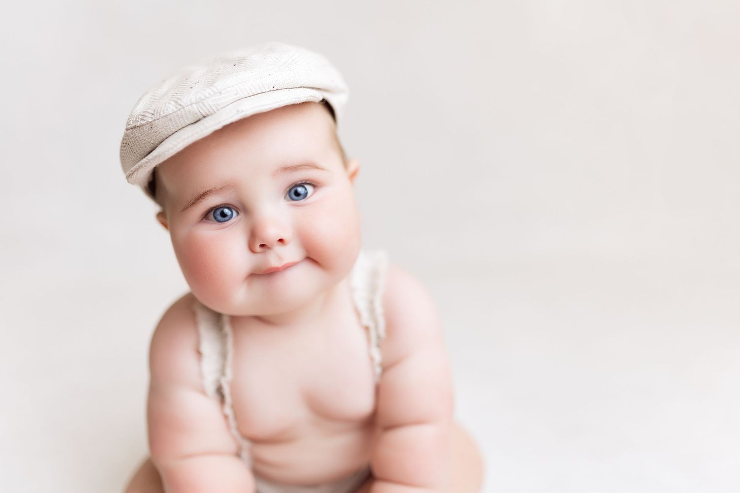 Sweet baby in newsboy cap sits upright against simple white background.