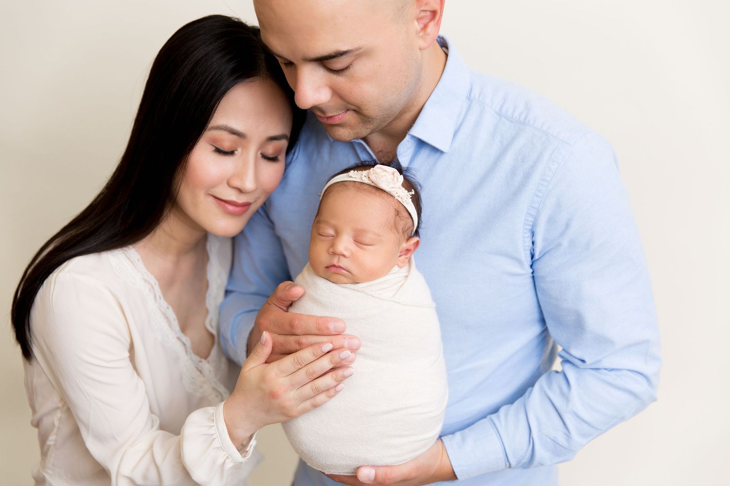 Family portrait with sleeping newborn wrapped in white being held by parents in soft lighting.
