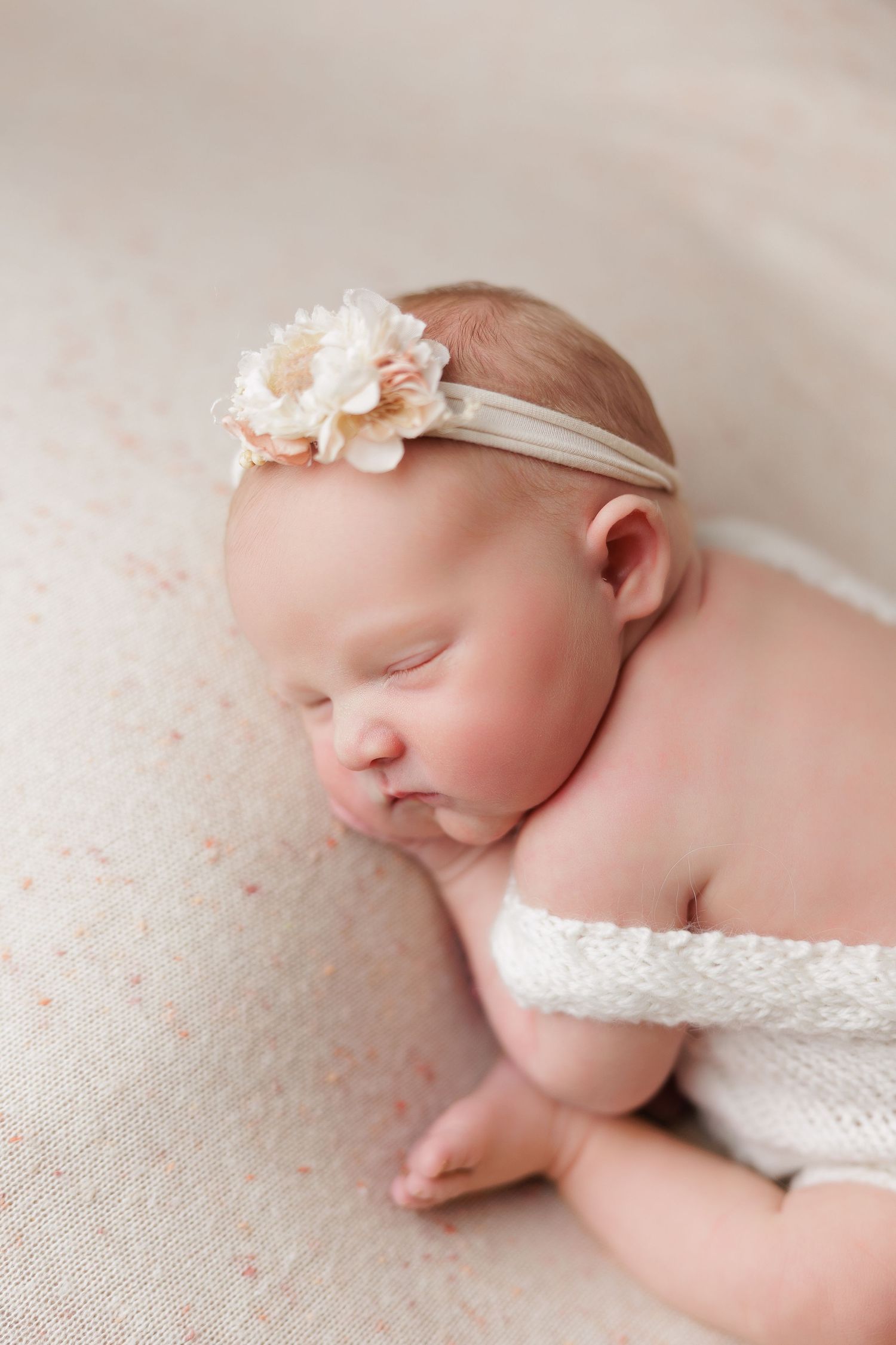 Sleeping newborn baby girl wearing a white floral headband and lace wrap, lying on a soft cream blanket.
