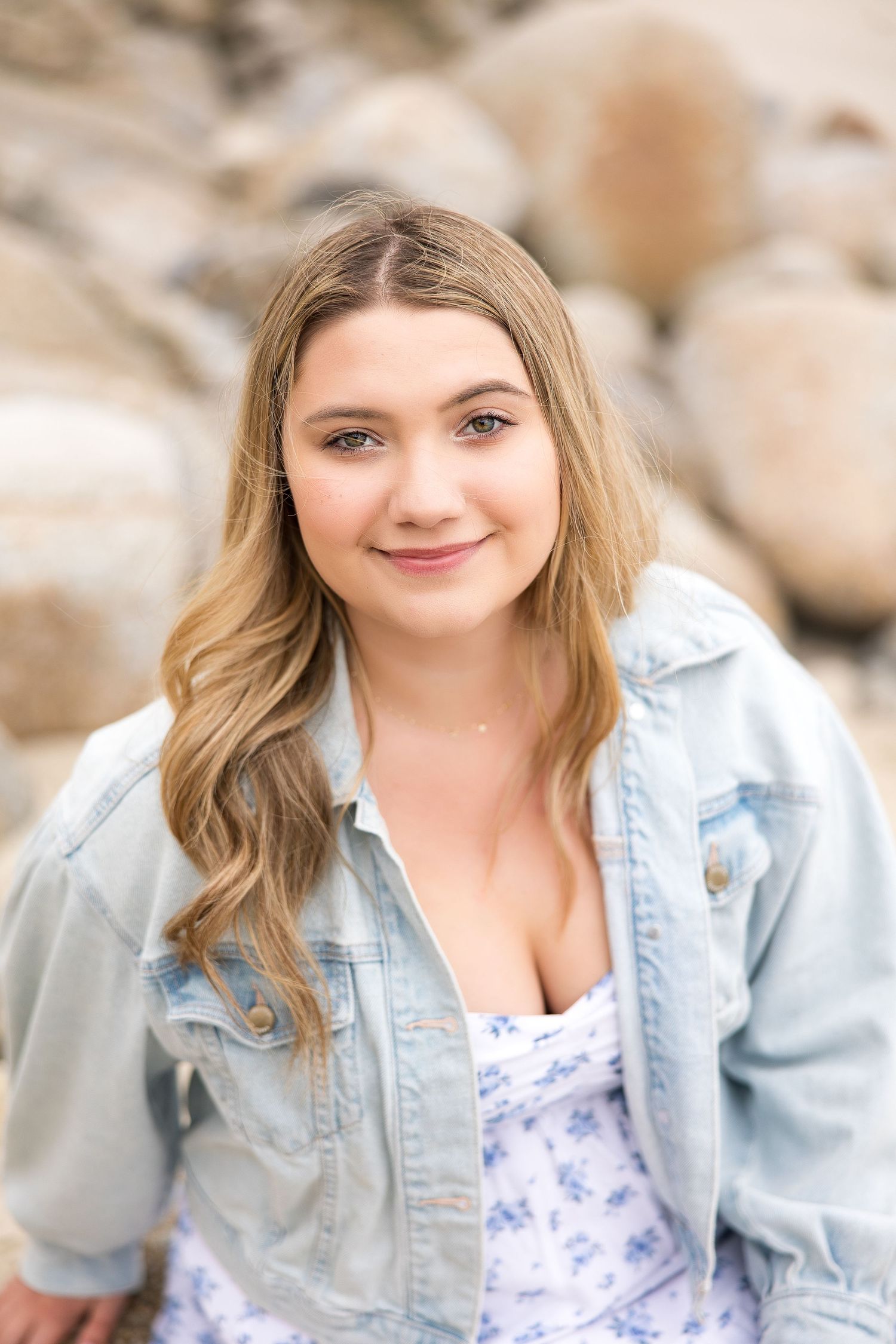 Young woman with wavy blonde hair smiling outdoors, wearing a denim jacket over a floral top, with rocks in background.