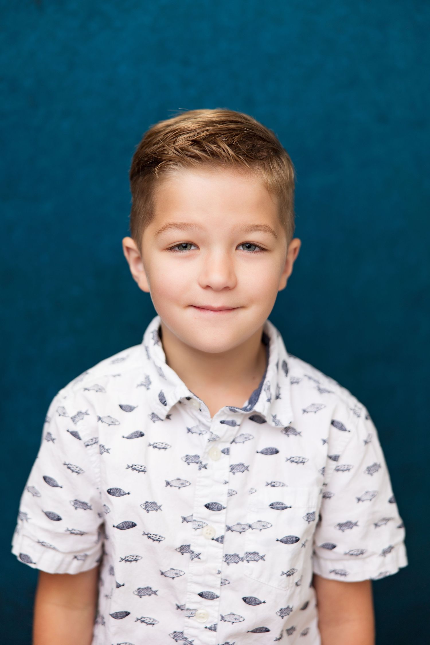 A young boy with blond hair smiles softly, wearing a white fish-print shirt against a teal background.