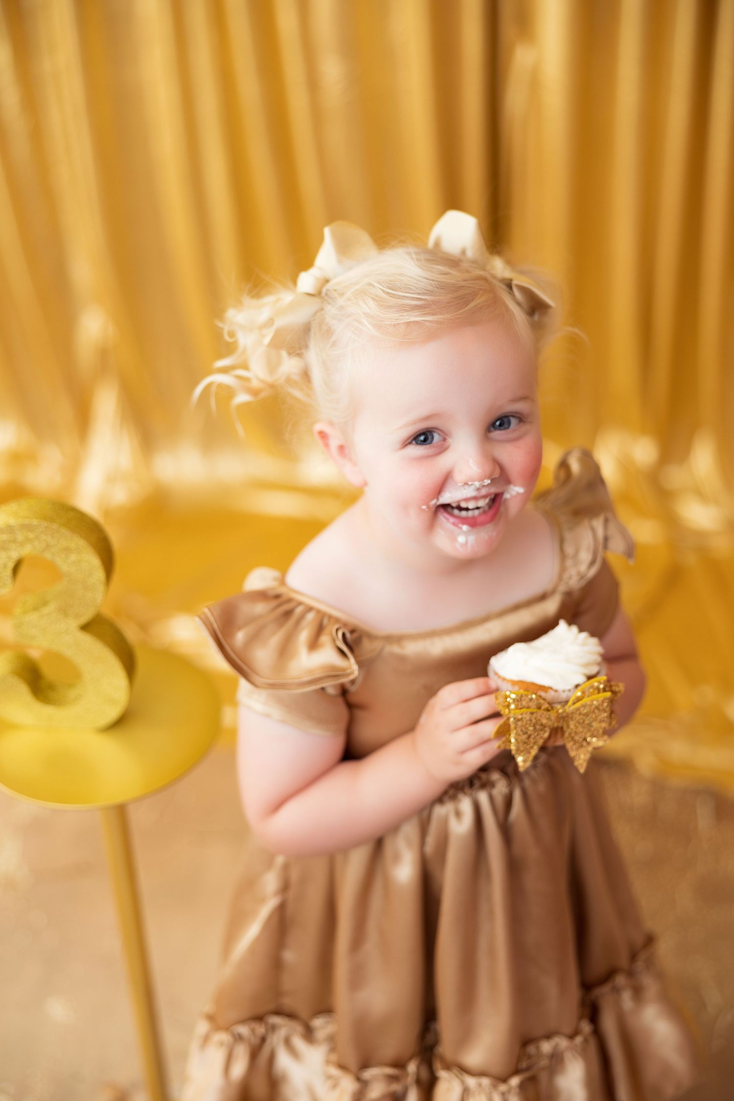 A toddler in a gold party dress laughs joyfully while holding a cupcake against a golden backdrop.