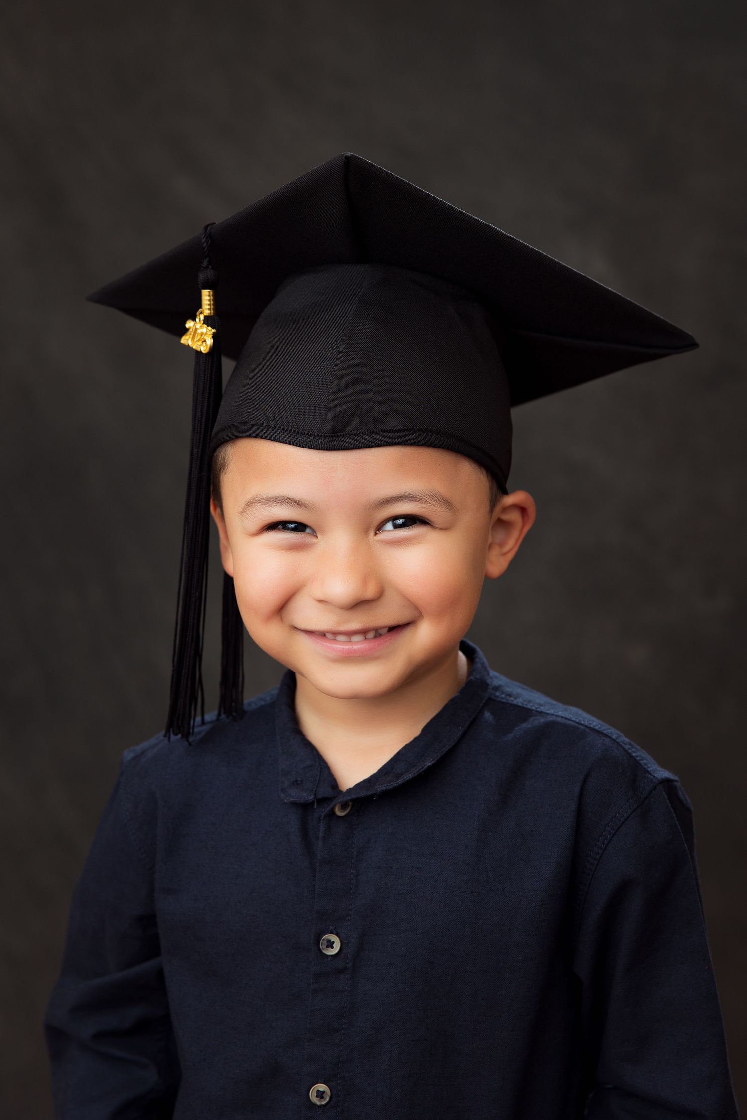 Young preschool graduate wearing navy blue gown and black mortarboard cap against dark background in portrait series.