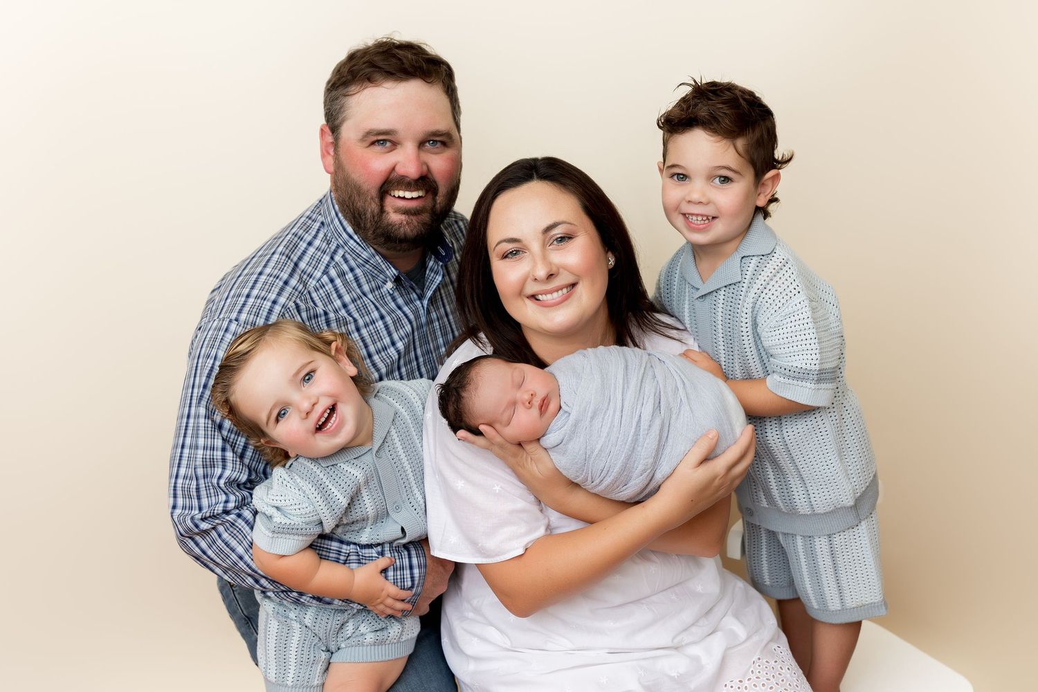 Family portrait against beige background showing parents with three children including a newborn in casual coordinated outfits.