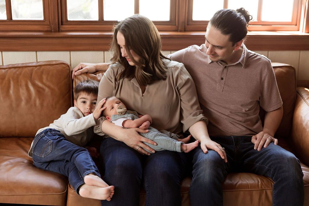 Family relaxes together on brown leather couch in cozy living room with natural window light.