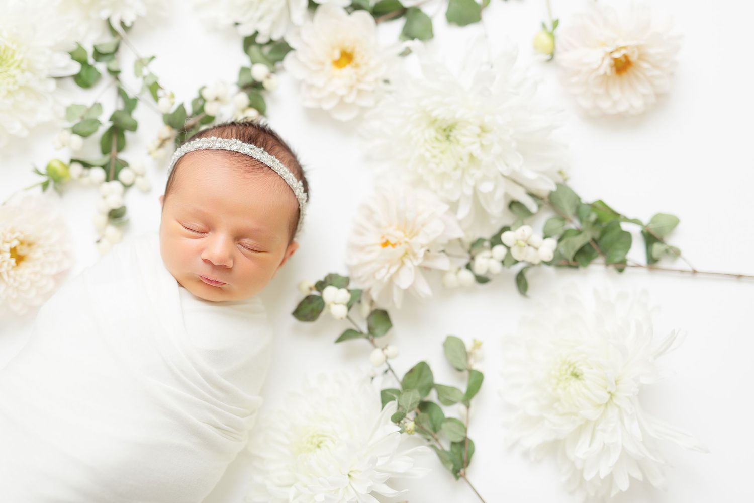 A sleeping newborn wrapped in white swaddle lies among delicate white dahlias and eucalyptus leaves on white backdrop.