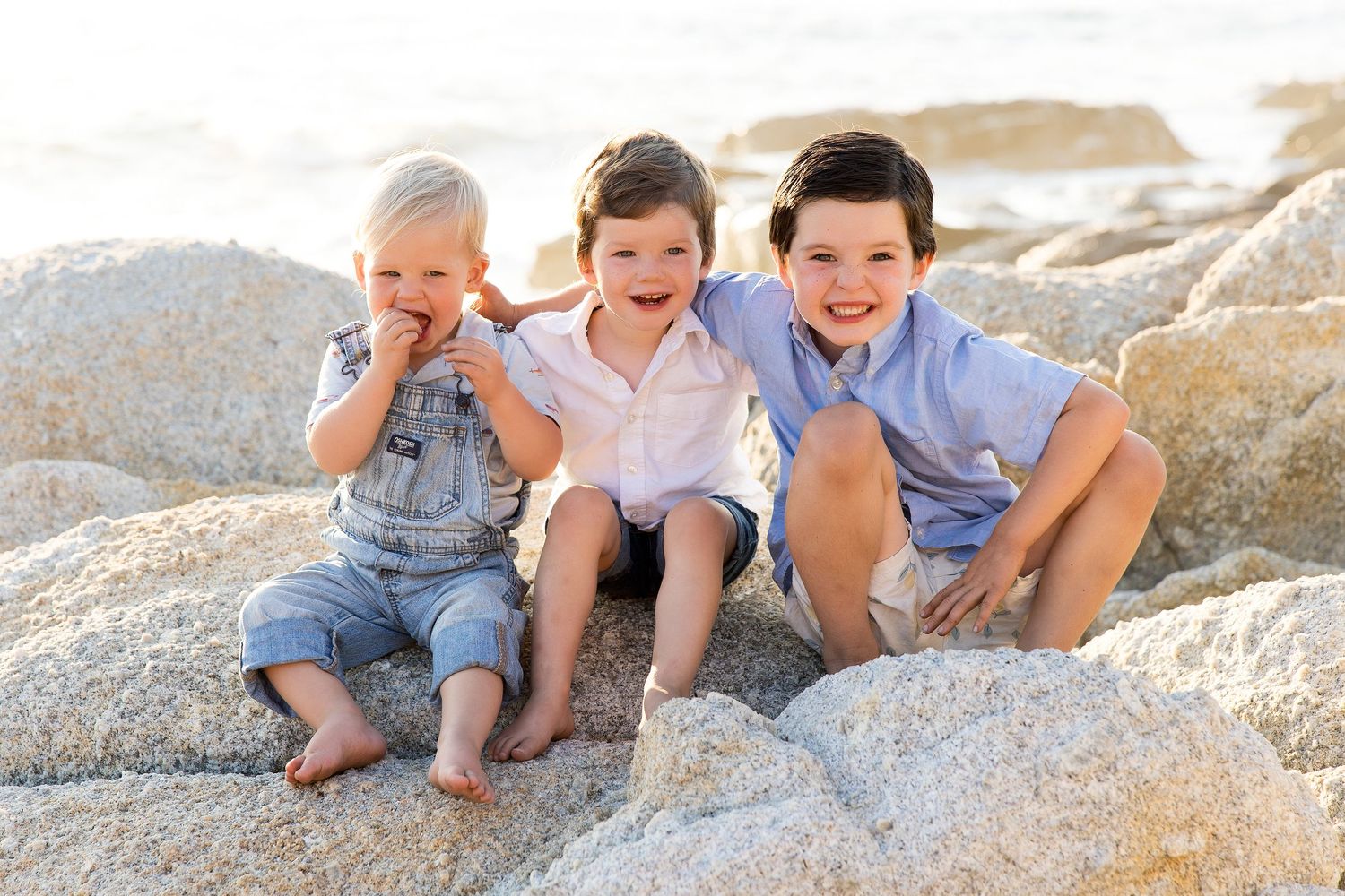 Three children sitting and laughing together on large beach rocks.