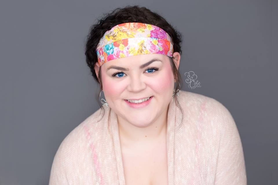 Smiling individual wearing a floral headband and light pink cardigan against a grey background.