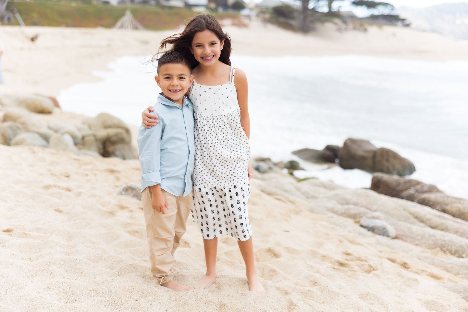 Children pose together on sandy beach with rocky coastline in background.