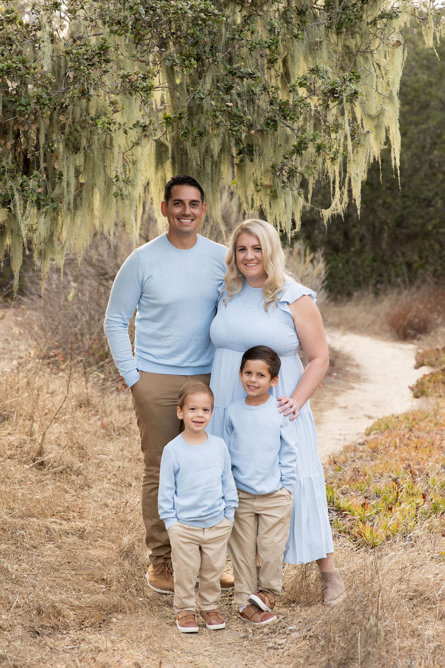 Family of four in coordinating light blue outfits pose together under moss-draped trees.