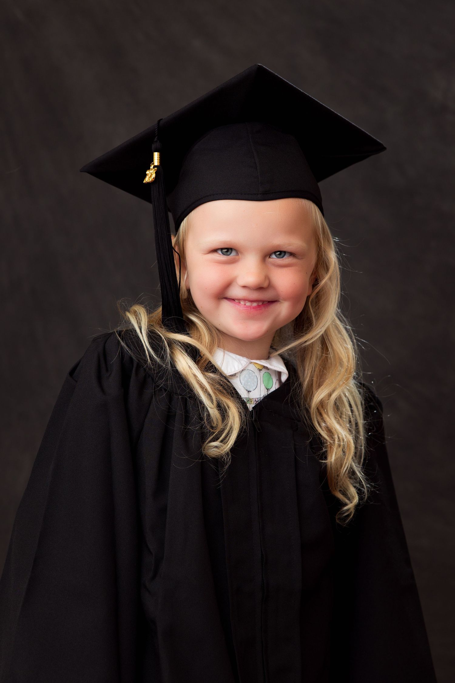Portrait of a young graduate wearing a black cap and gown with blonde hair against a dark background.