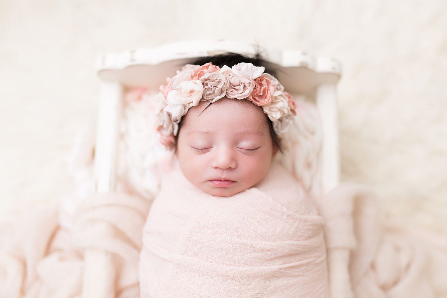 A sleeping newborn wrapped in soft pink swaddle blanket wears a delicate floral headband while resting peacefully.