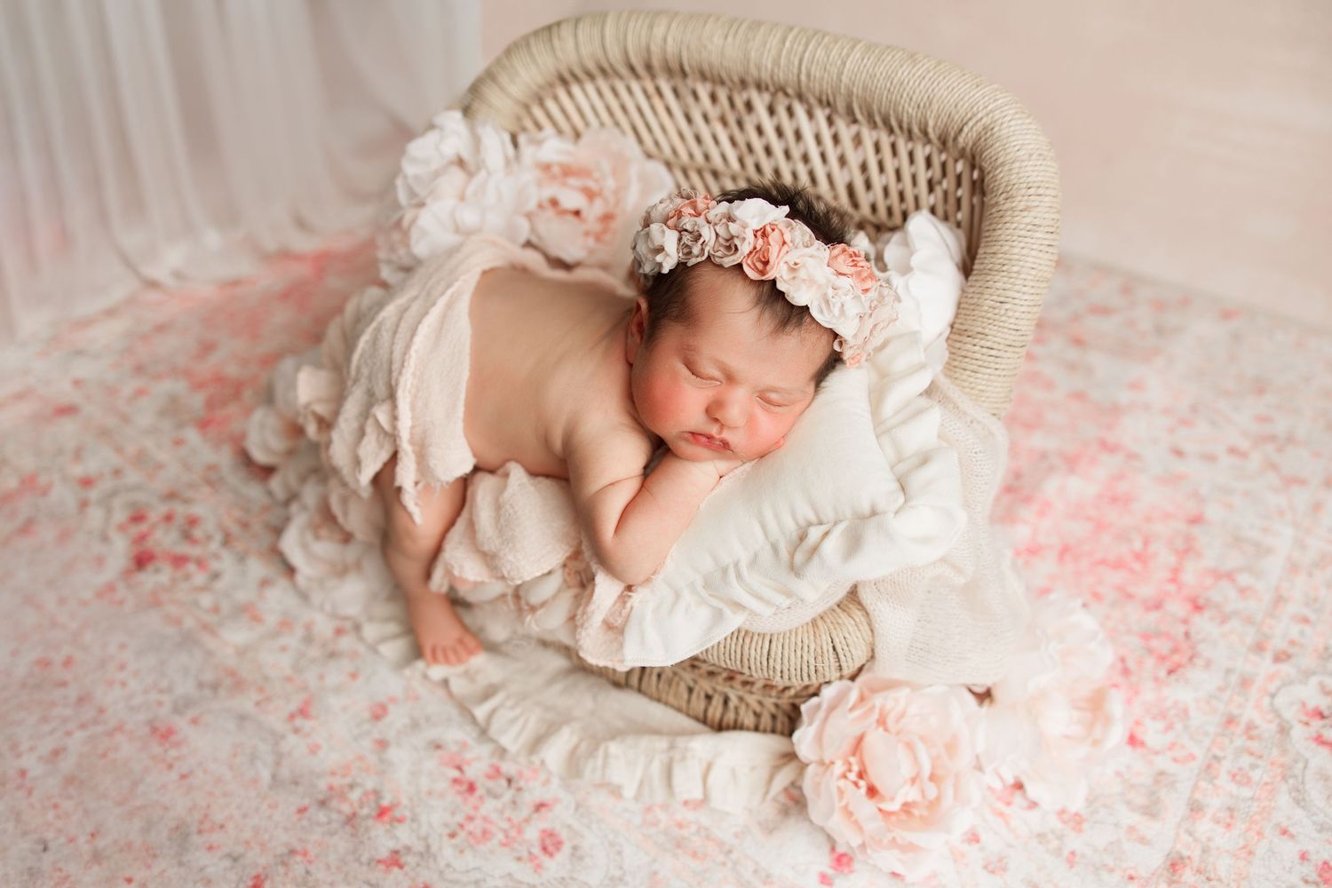 Sleeping newborn in wicker basket with pink flowers and lace headband on floral blanket.