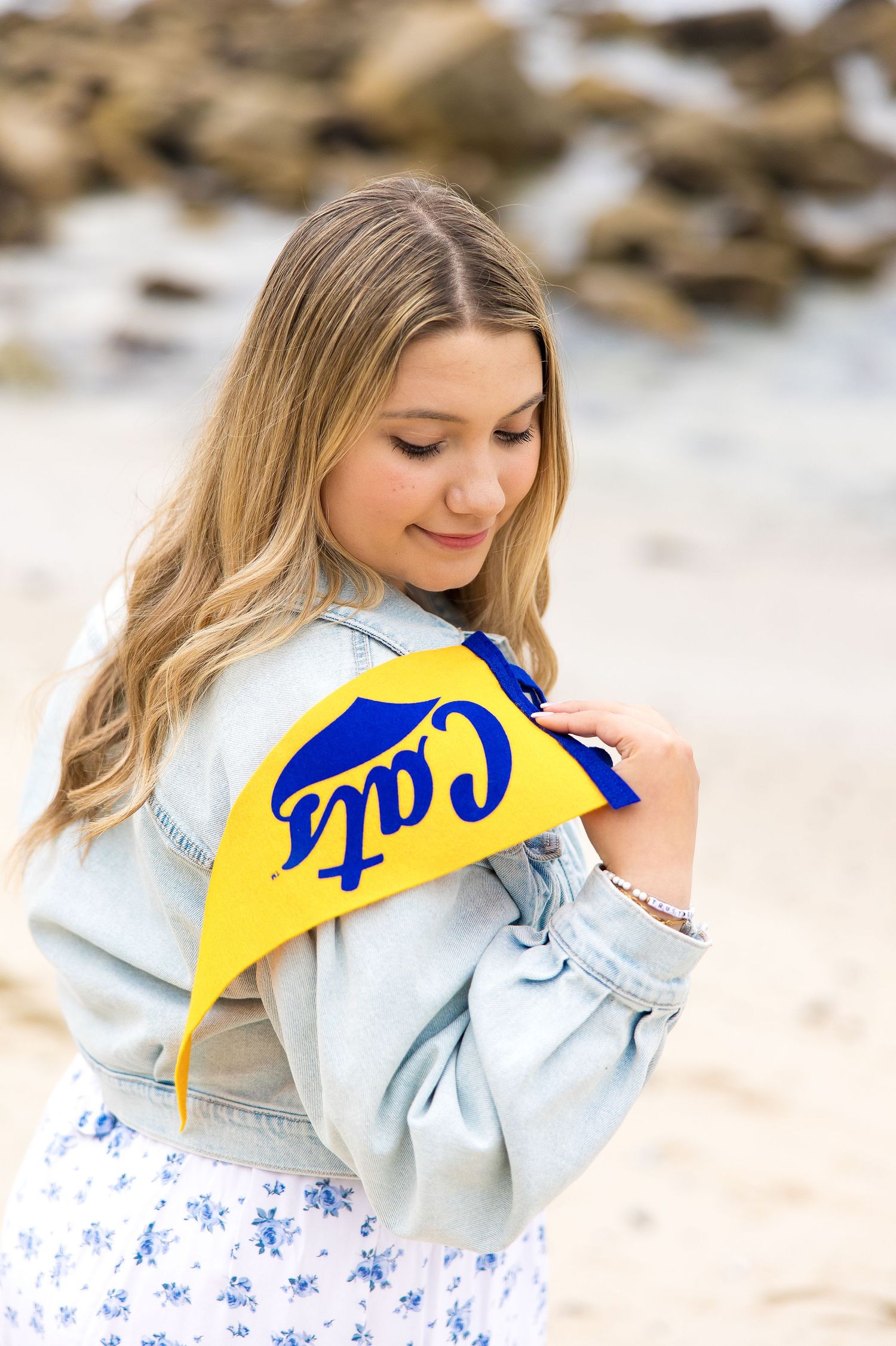 Young woman in denim jacket holding yellow and blue Cats pennant flag at a rocky beach.