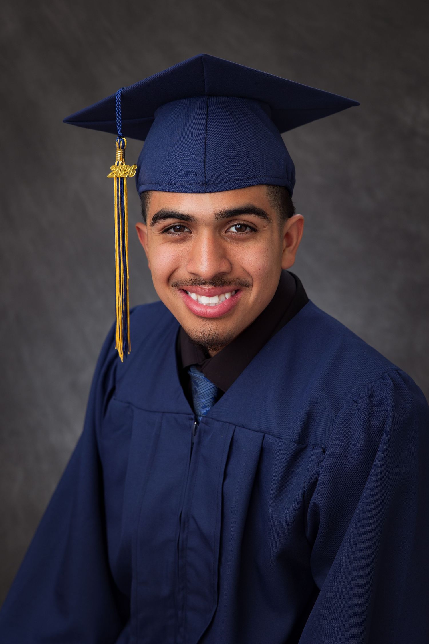 Smiling young man in navy blue graduation cap and gown with gold tassel posing for senior portrait.