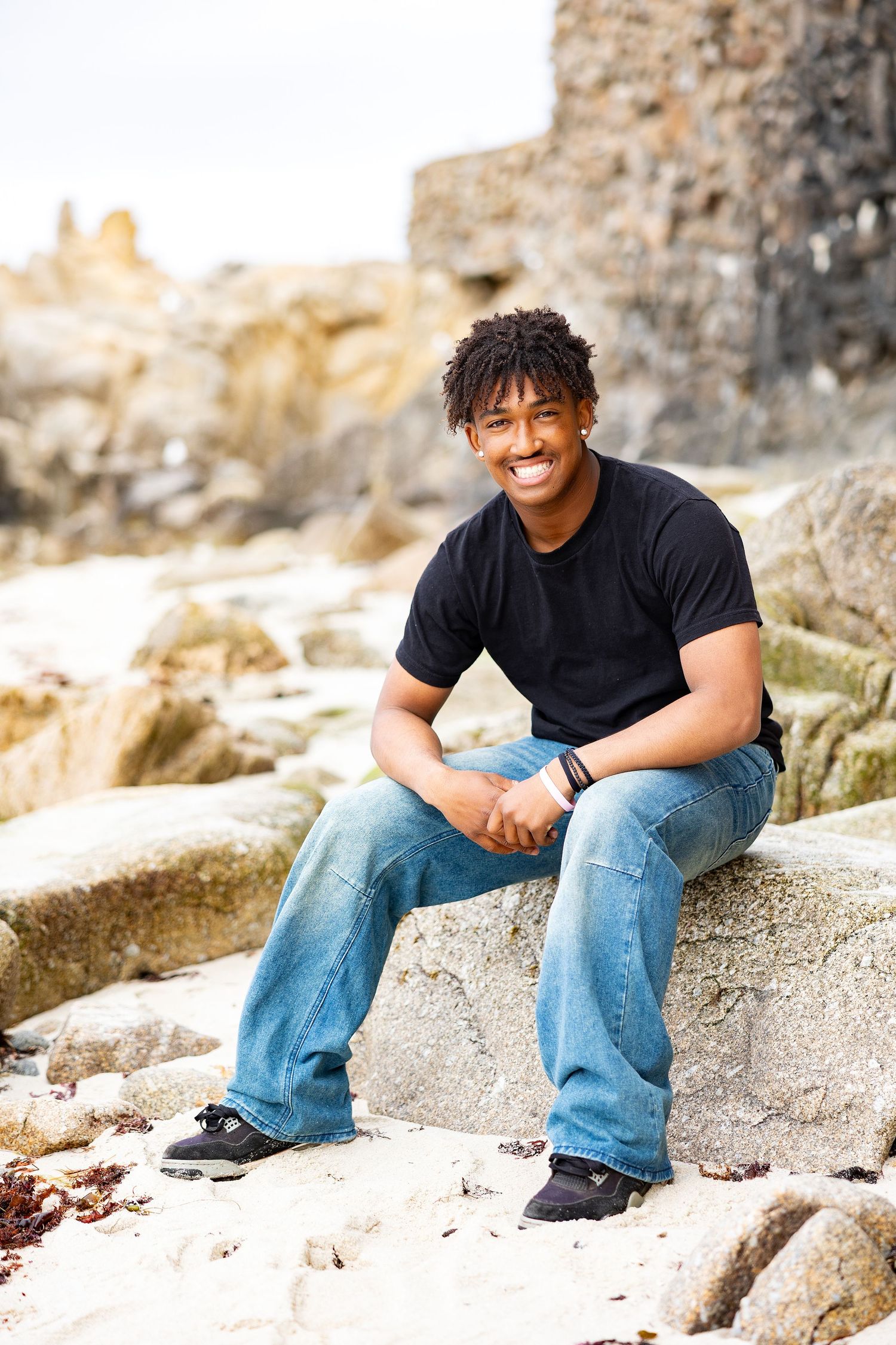 Smiling young Black man sitting on rocks at a beach wearing jeans and a black t-shirt.