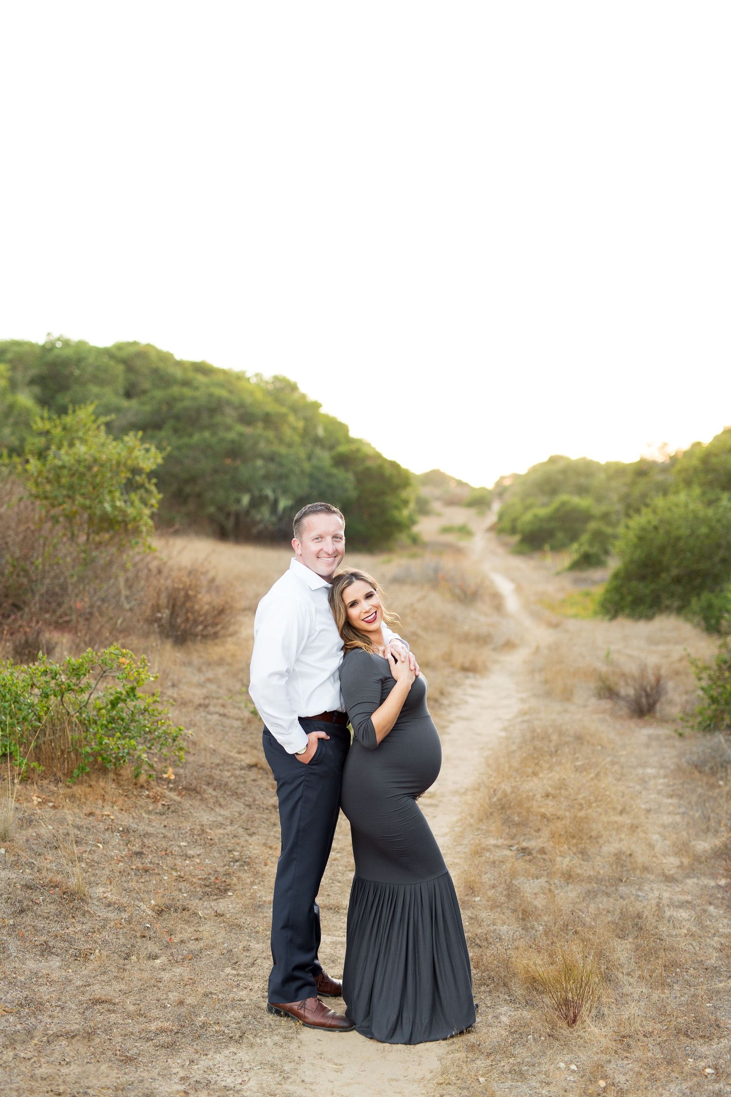 A couple embraces tenderly on a winding dirt path surrounded by coastal brush and golden light.