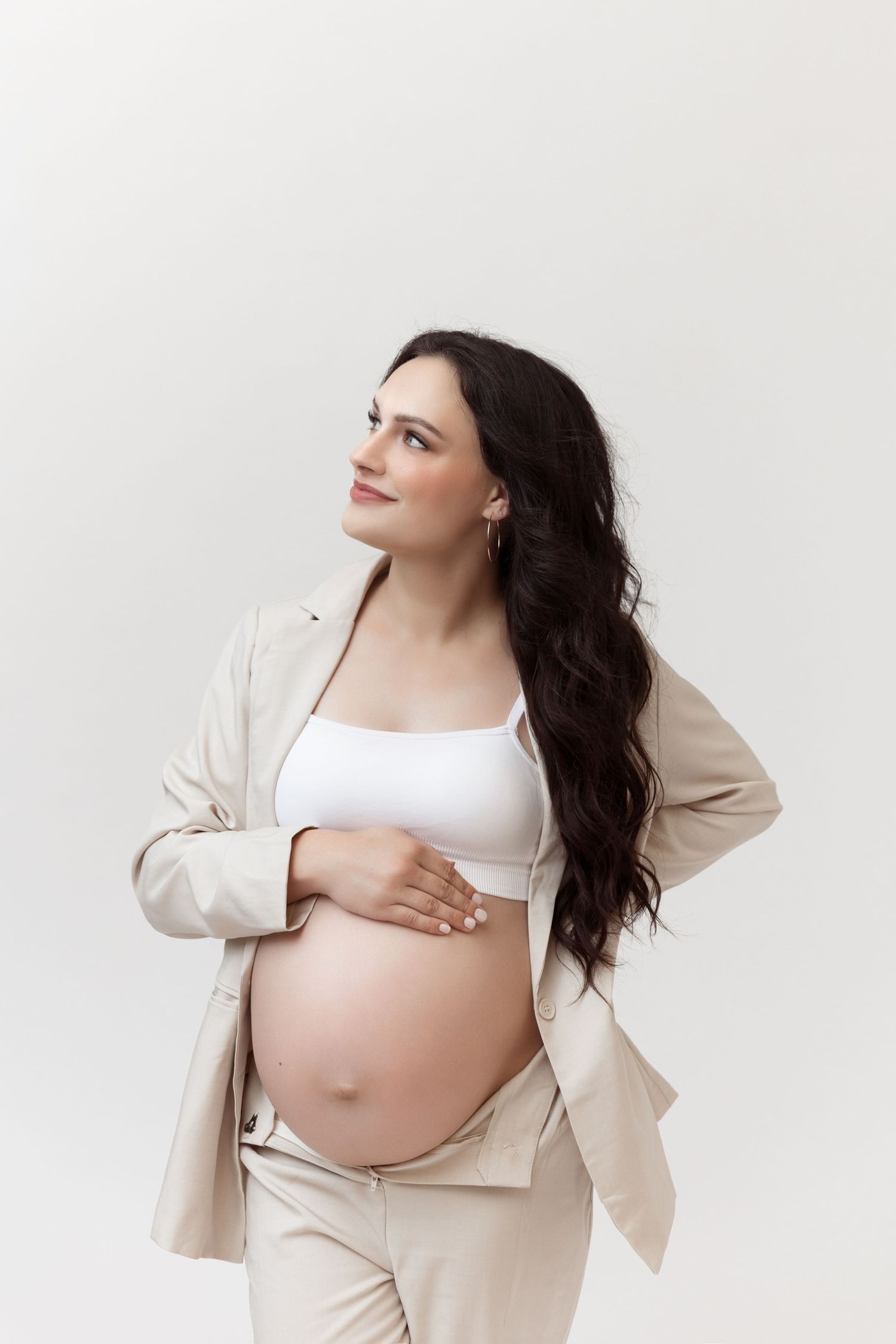 Maternity photoshoot featuring woman in beige blazer and white top posing against light background.