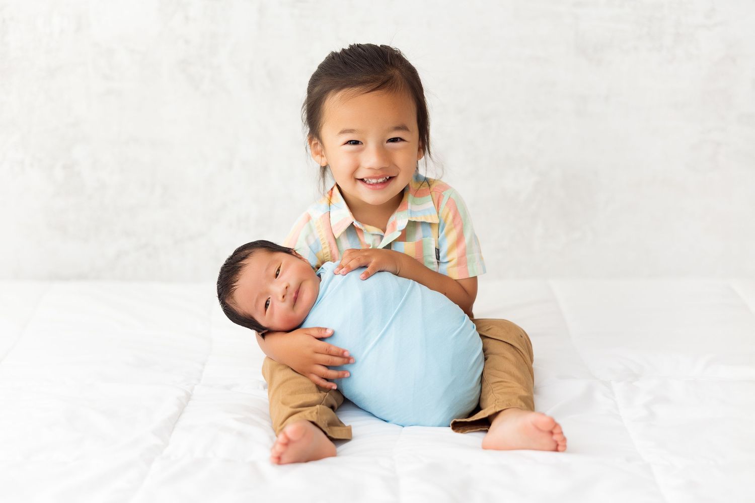 Two siblings posing on white backdrop, older child holding younger baby in blue outfit.