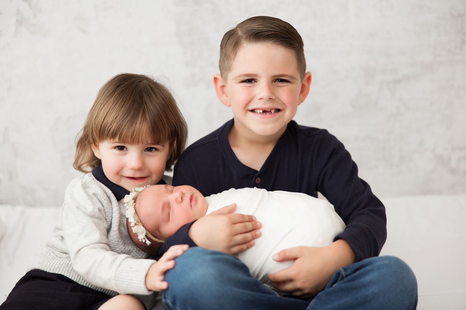 Three siblings posing together with newborn baby against light backdrop.