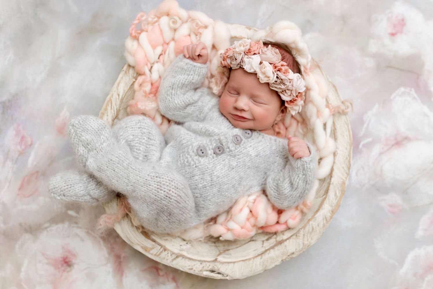Sleeping newborn in cozy basket wearing gray wrap and floral bonnet.