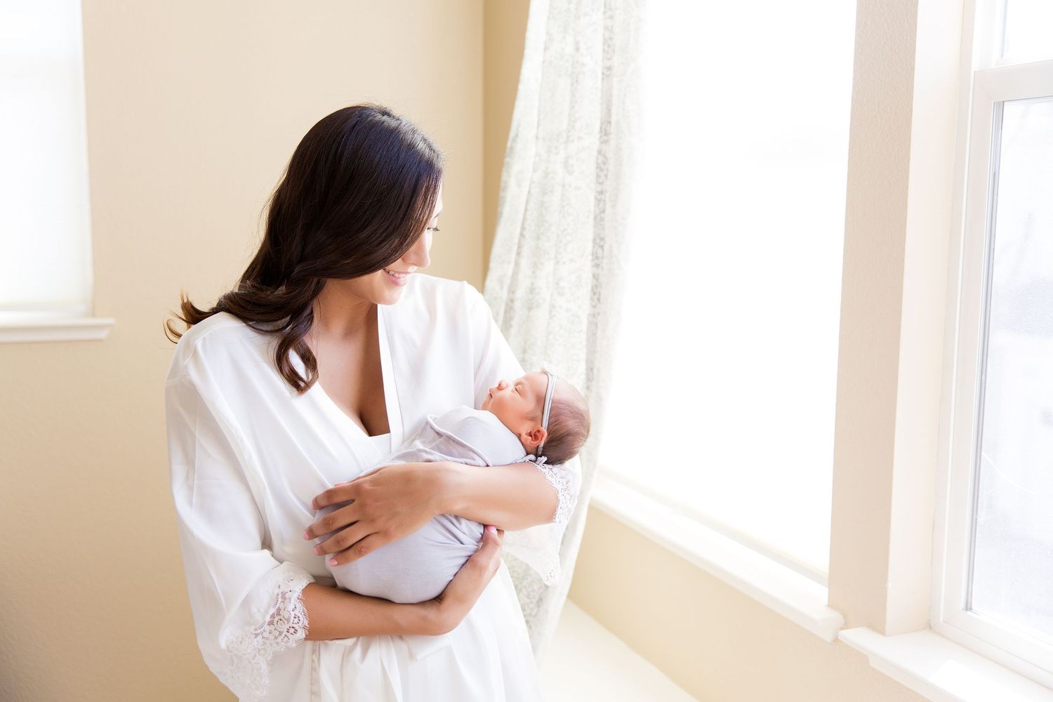 Mother in white robe cradles sleeping newborn baby near bright window.