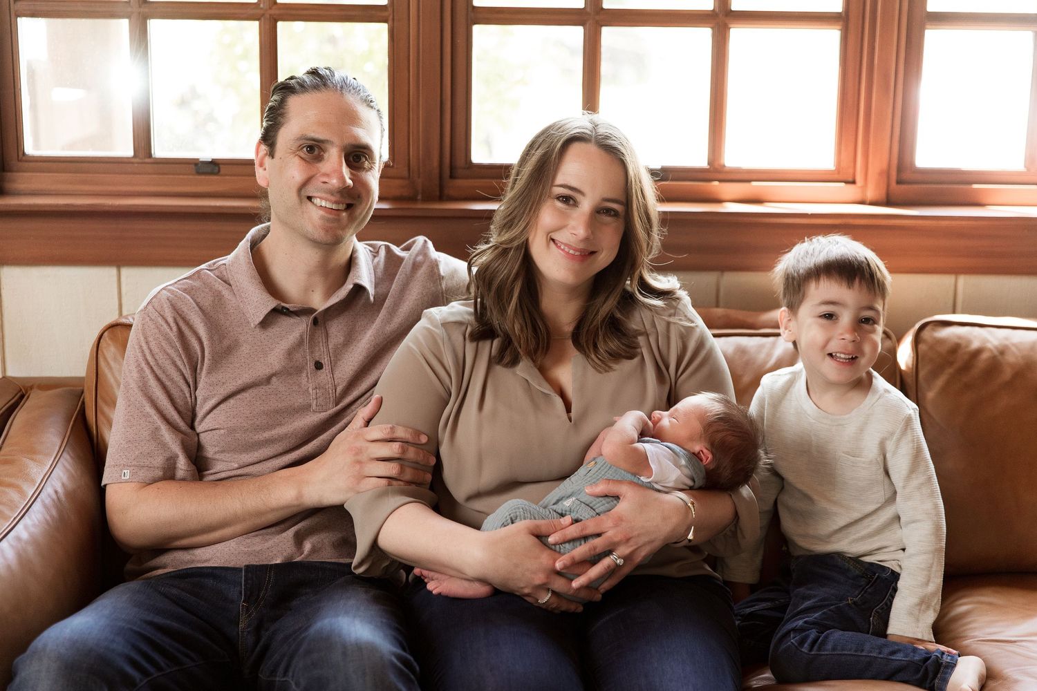 Family relaxing together on brown leather couch in sunlit room.
