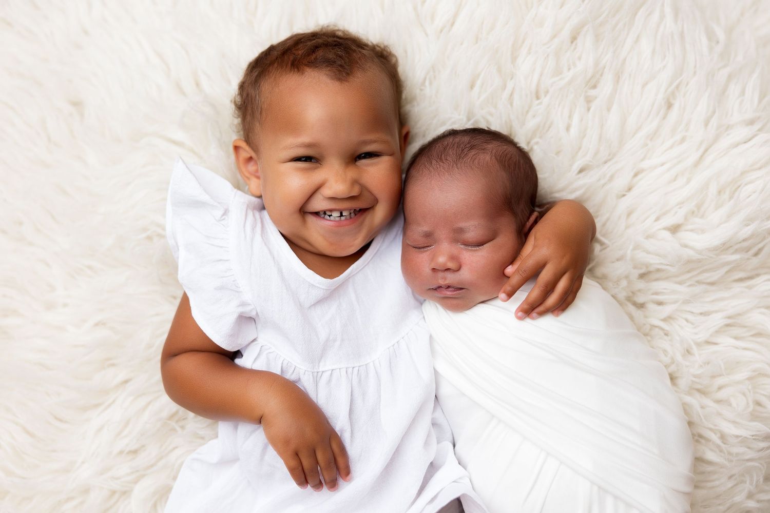 Two young children in white outfits pose together on a soft white blanket.