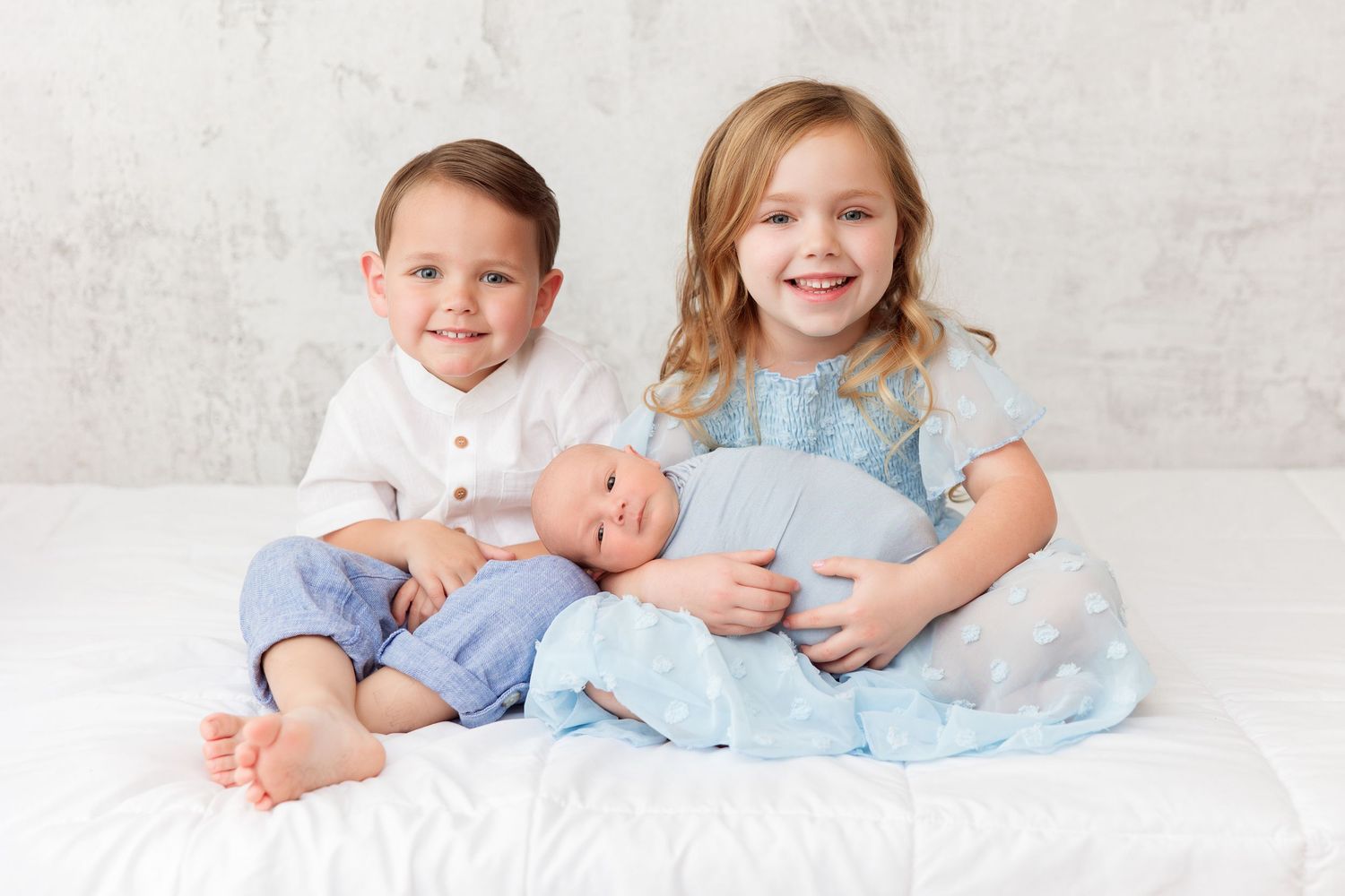 Children sitting together on white bed with baby sibling.