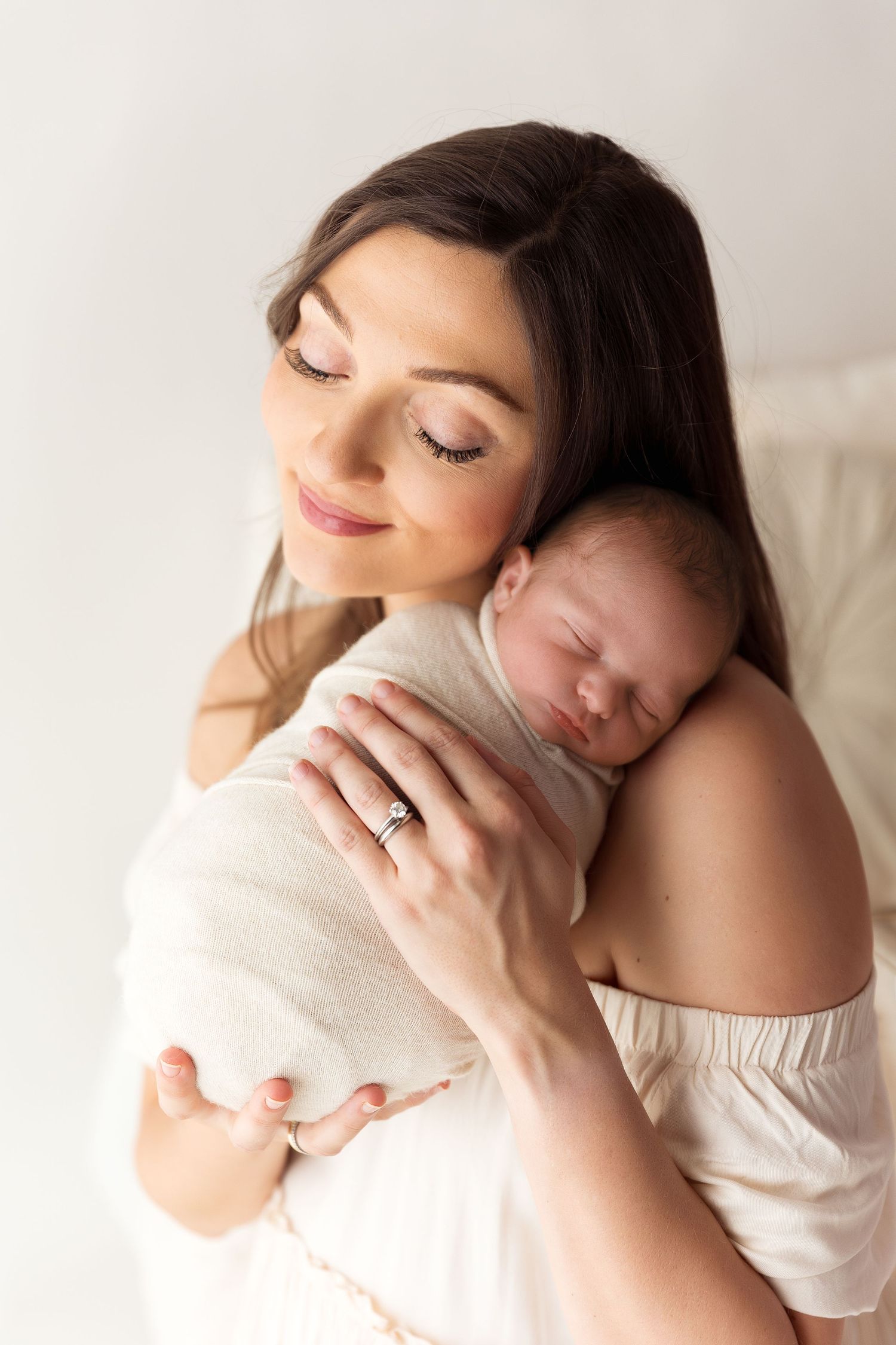 A tender moment as mother gently embraces her sleeping newborn baby against her shoulder in soft natural light.