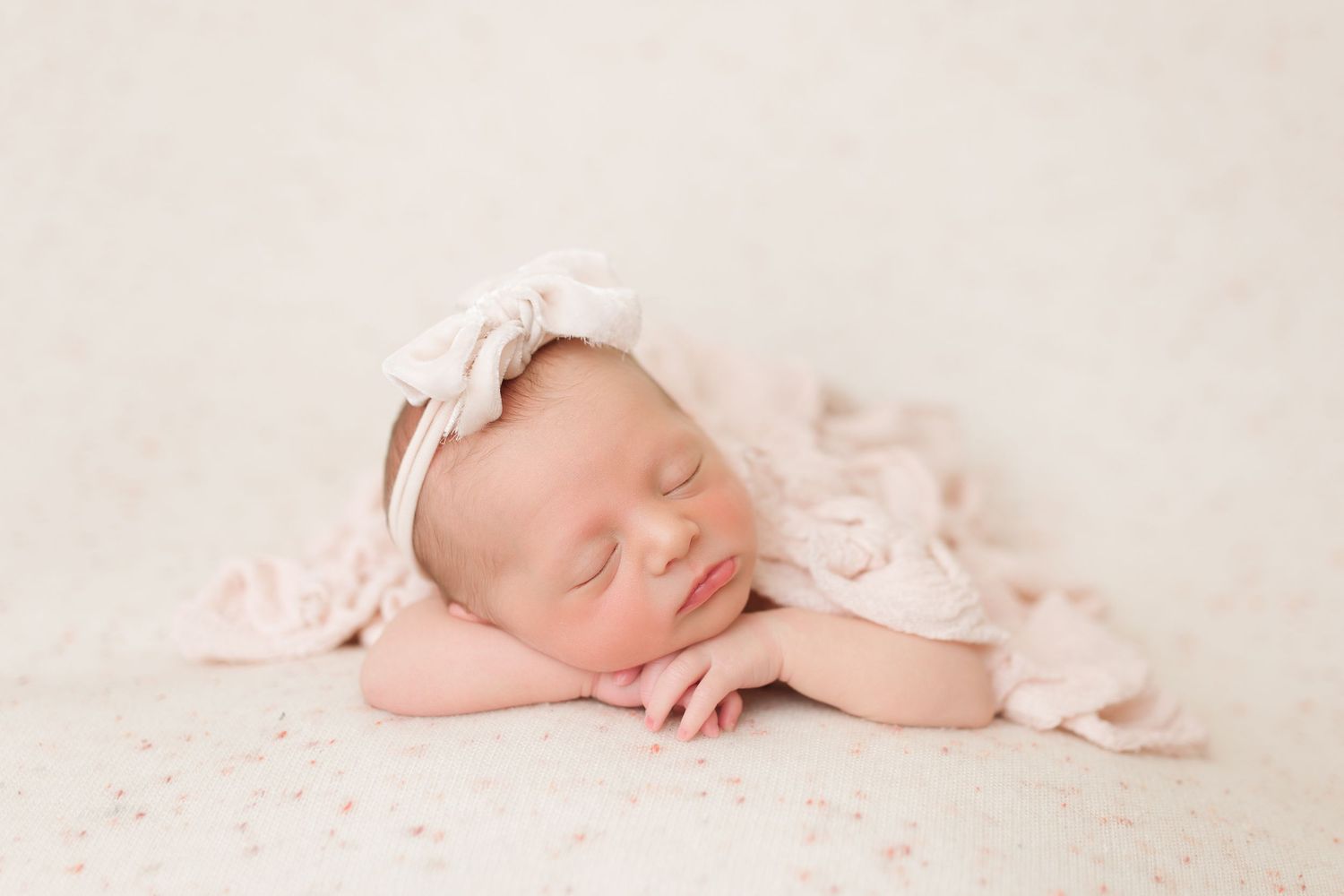Newborn baby sleeping peacefully under soft pink fabric with delicate headband.