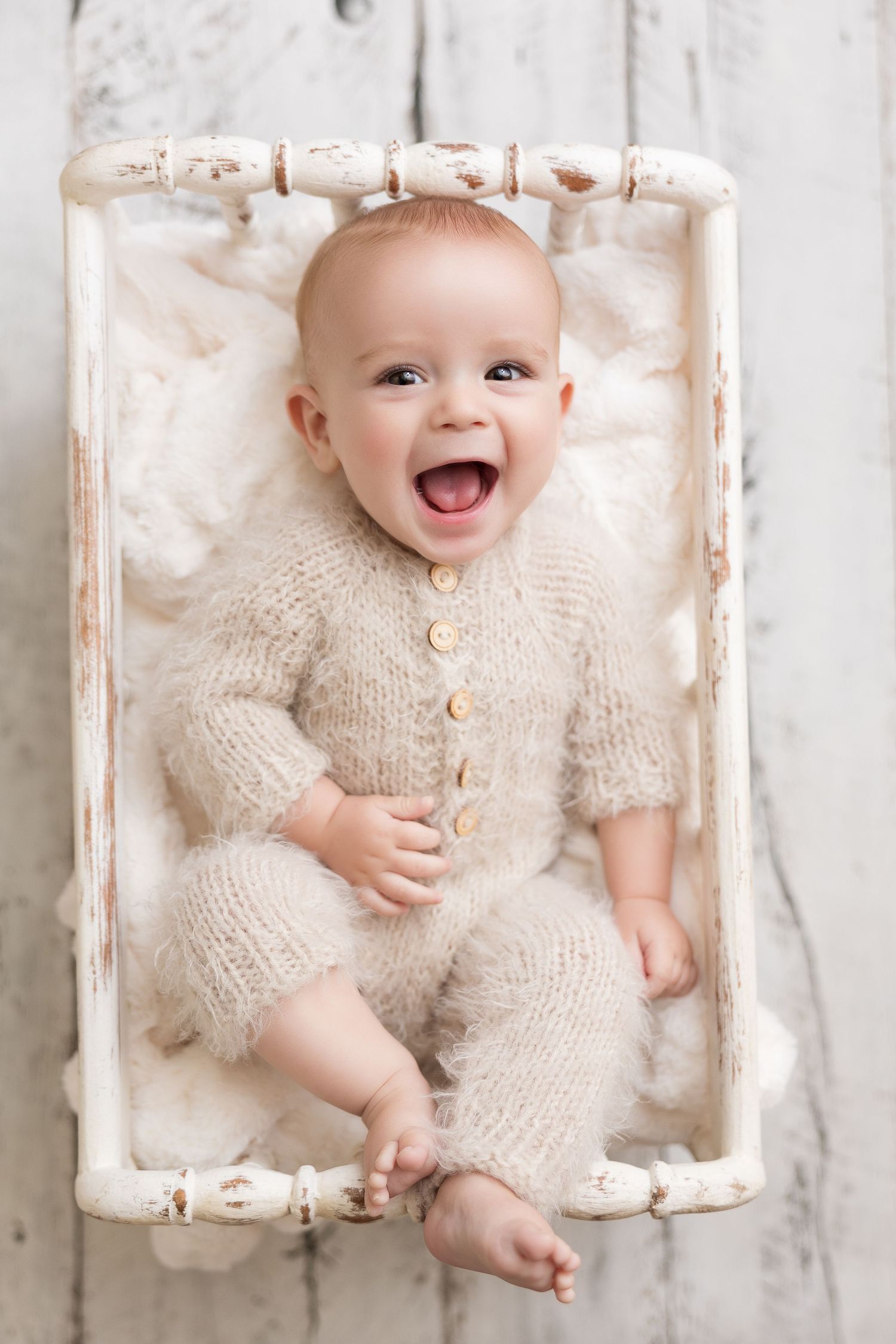 Baby wearing cream knit romper lies smiling on white textured backdrop during professional photo session.