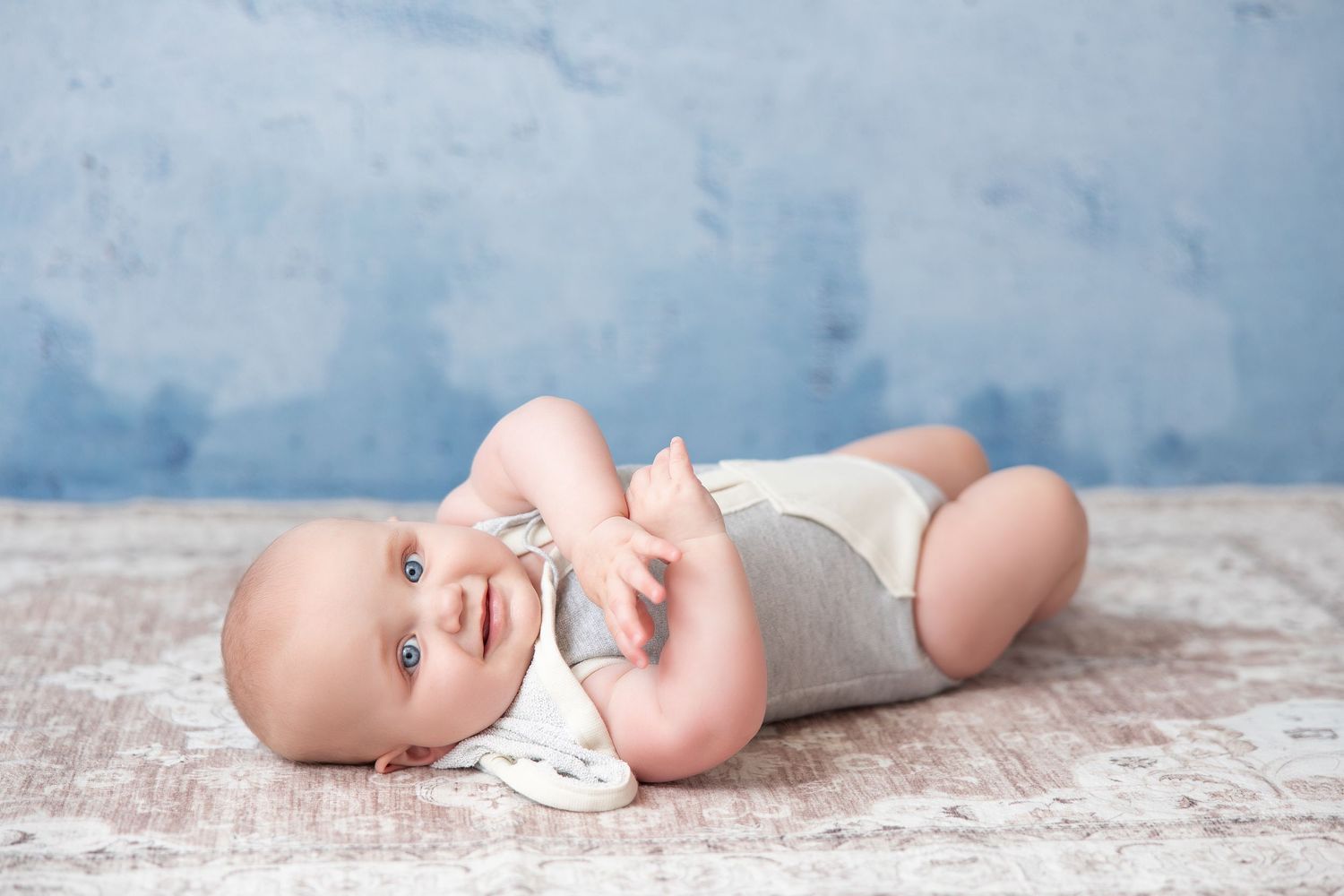 Baby in gray onesie lying on back against blue backdrop.