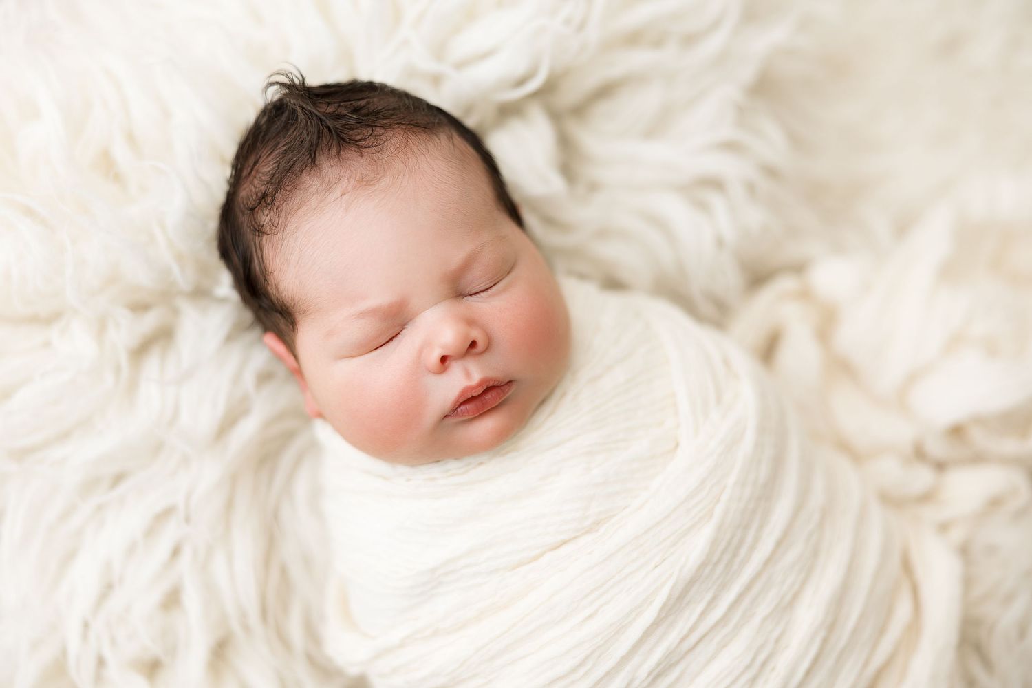 Sleeping newborn baby wrapped in soft white textured blanket during professional photo session.