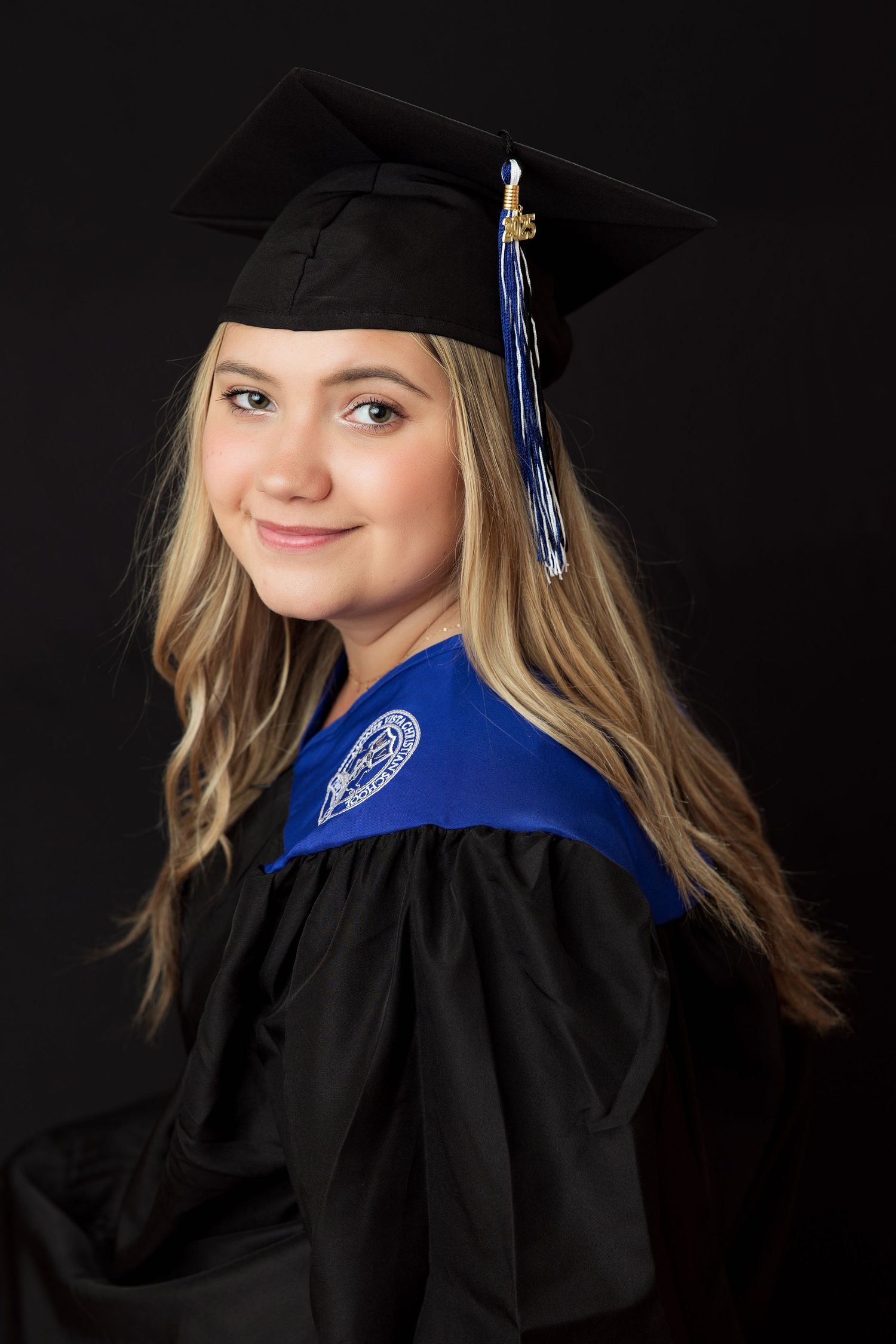Young woman in black graduation cap and gown with blue academic hood smiles over shoulder against dark background.