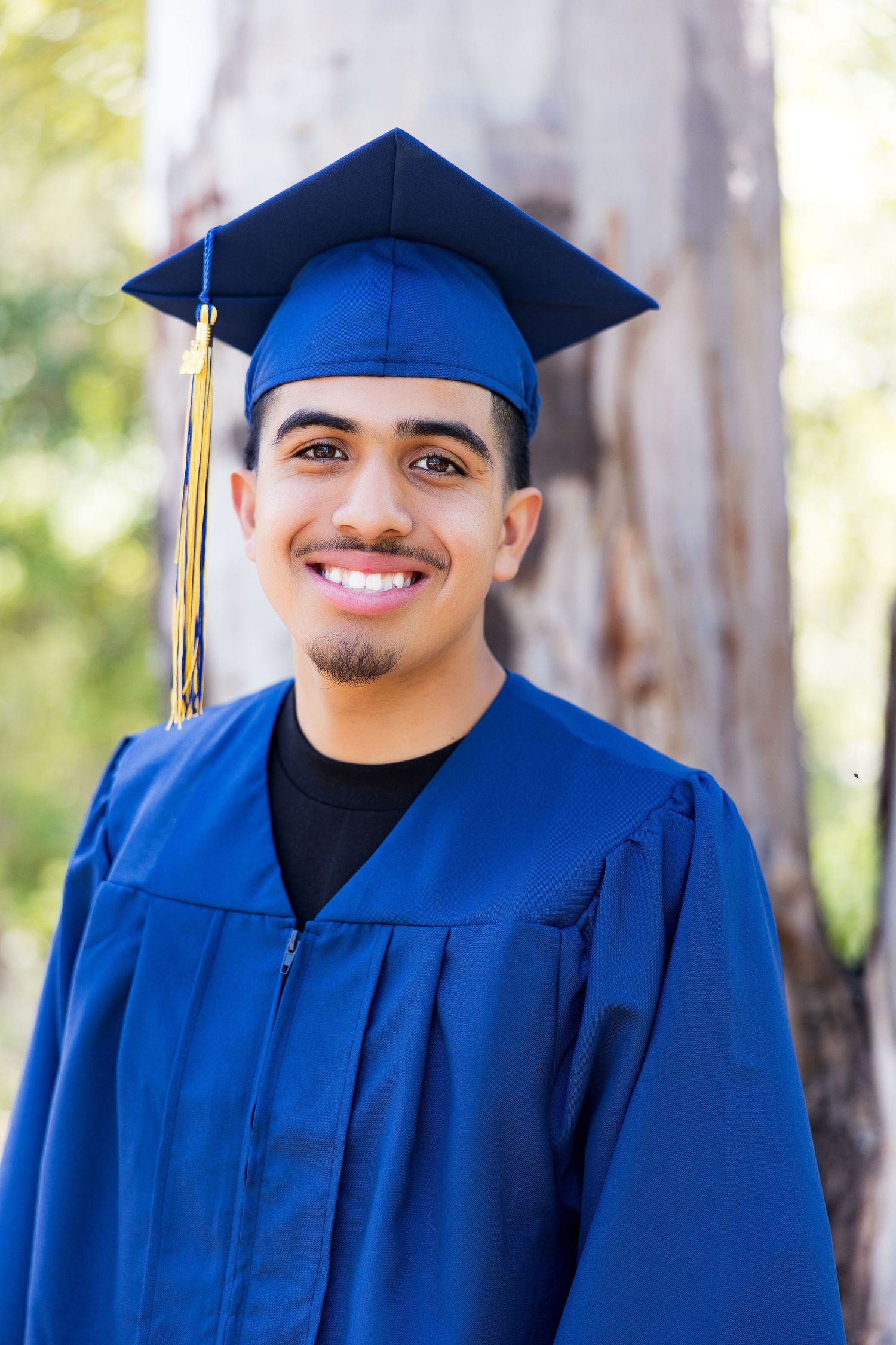 Young man smiling in blue graduation cap and gown outdoors, celebrating academic achievement.