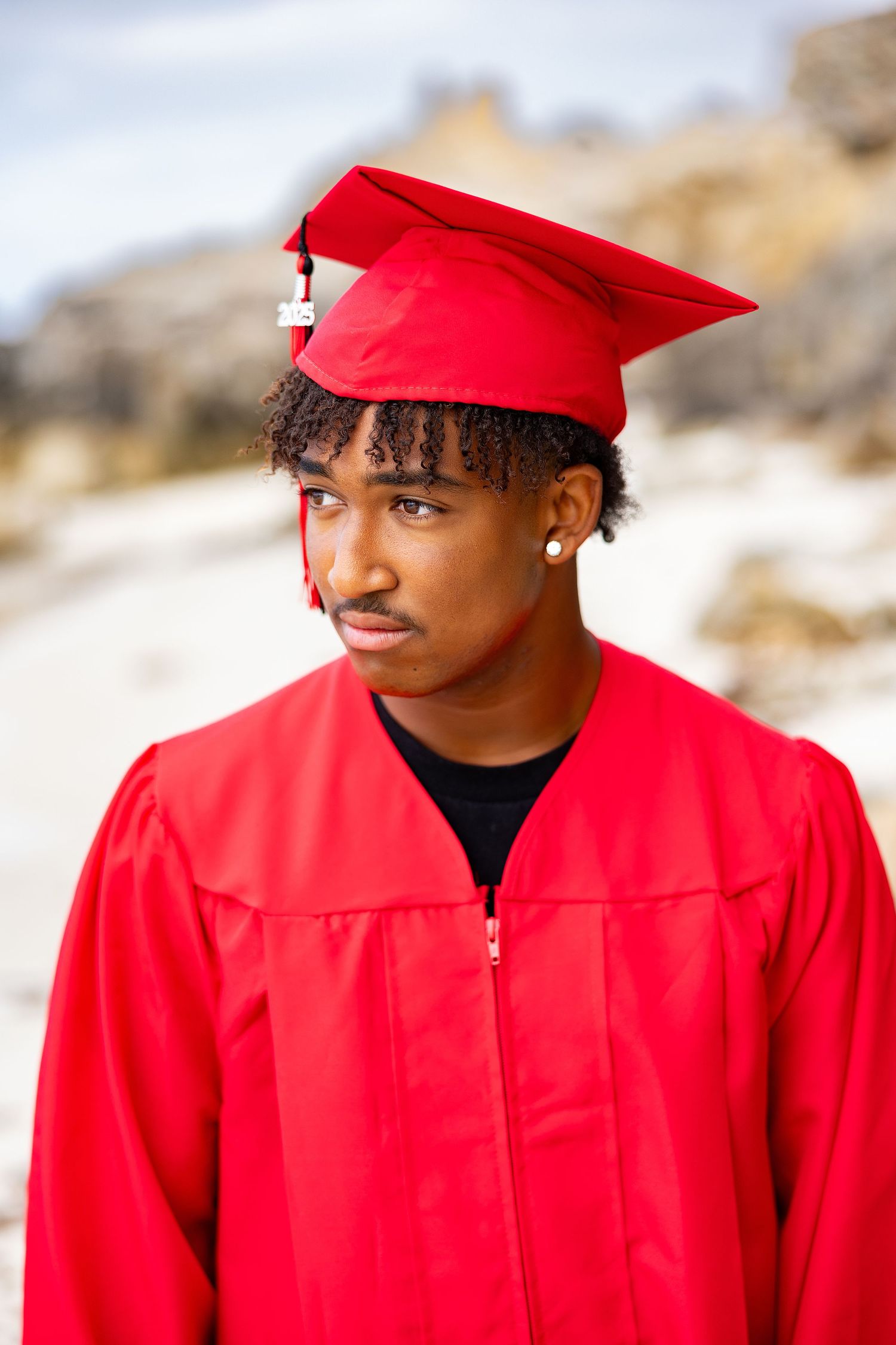 Young man in red graduation cap and gown standing outdoors looking to the side.