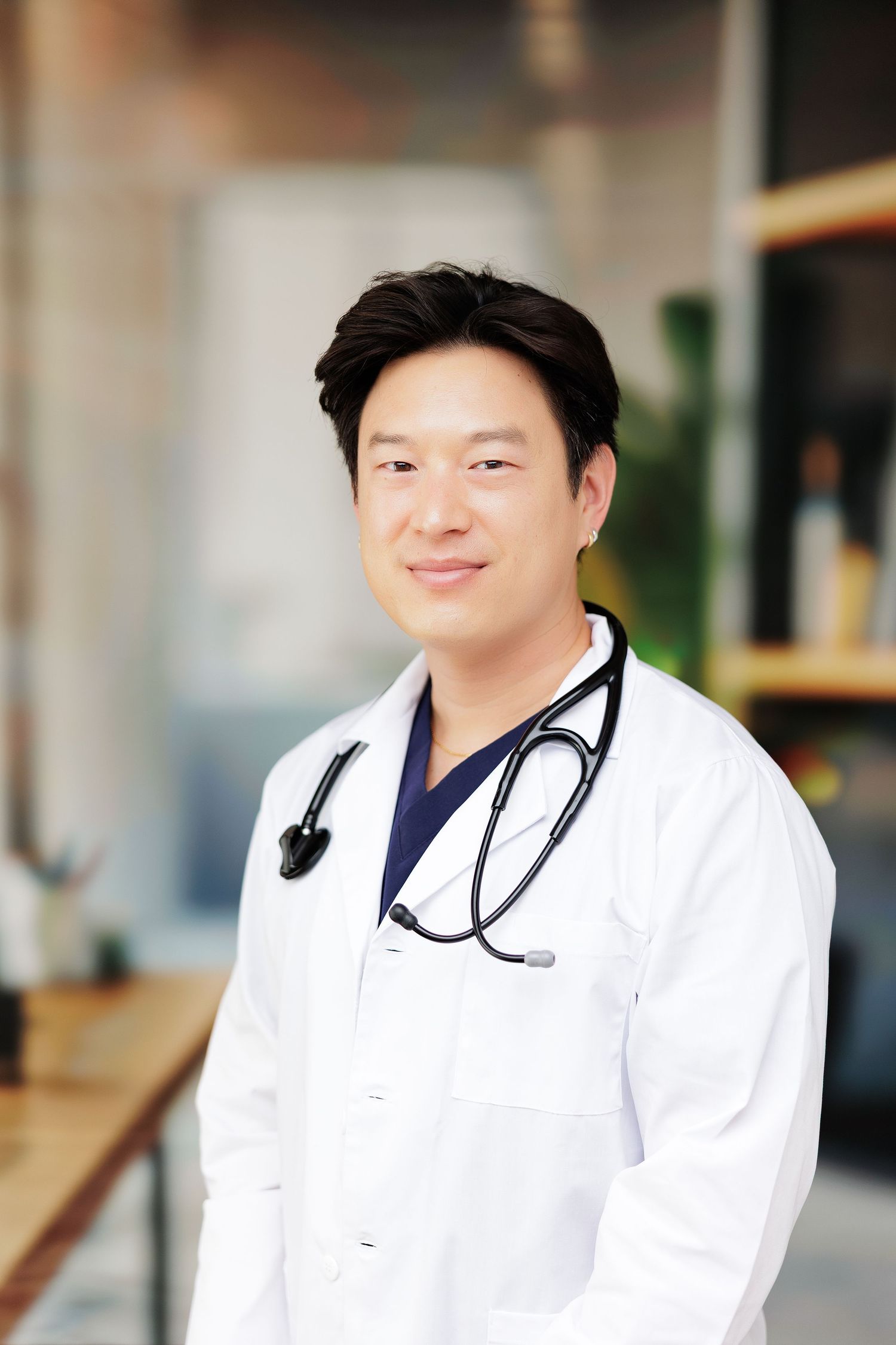 Asian male doctor in white lab coat with stethoscope around neck smiling confidently in modern medical office.