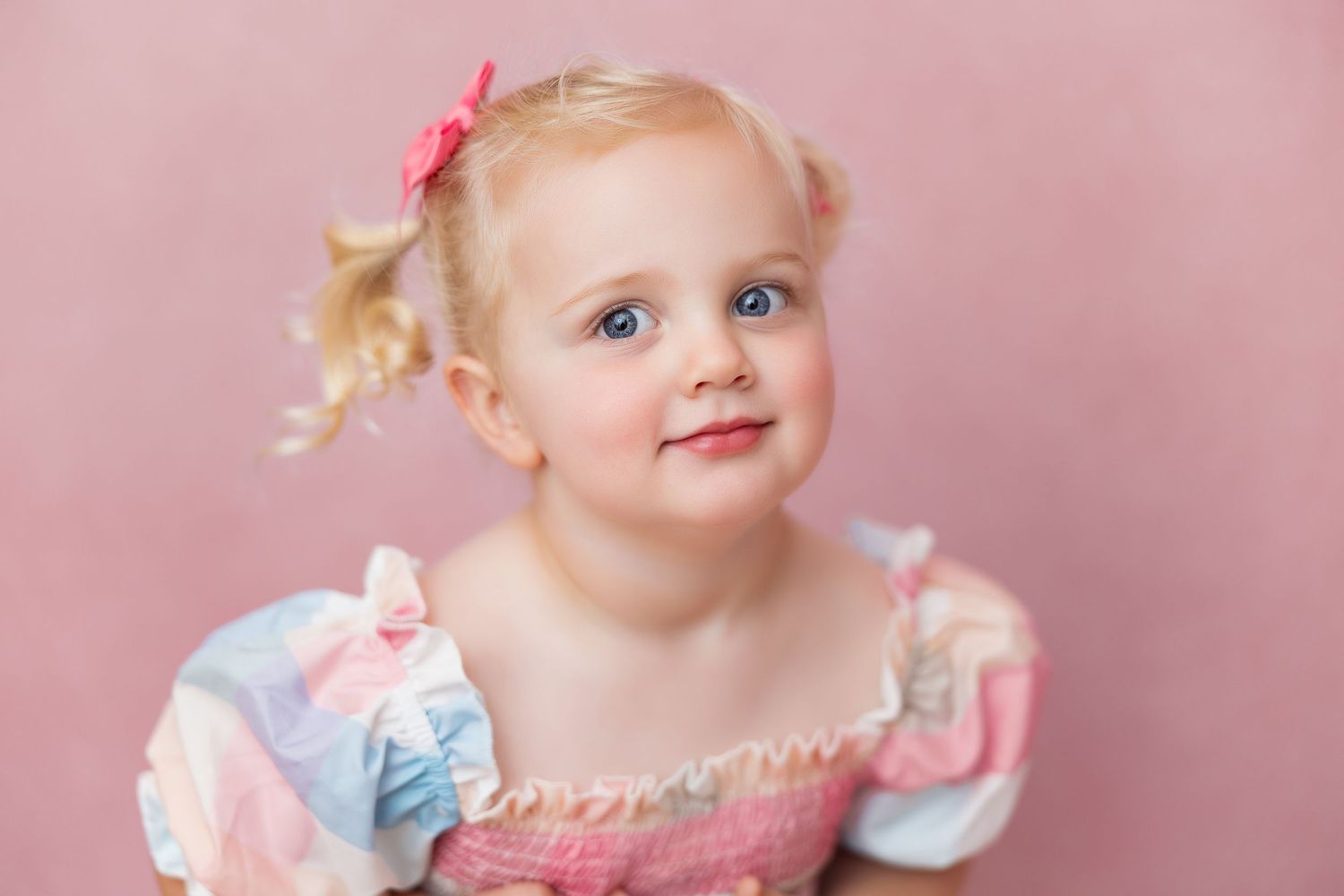 A baby wearing a light pink dress and blue bow sits in front of a soft pink backdrop during a portrait session.