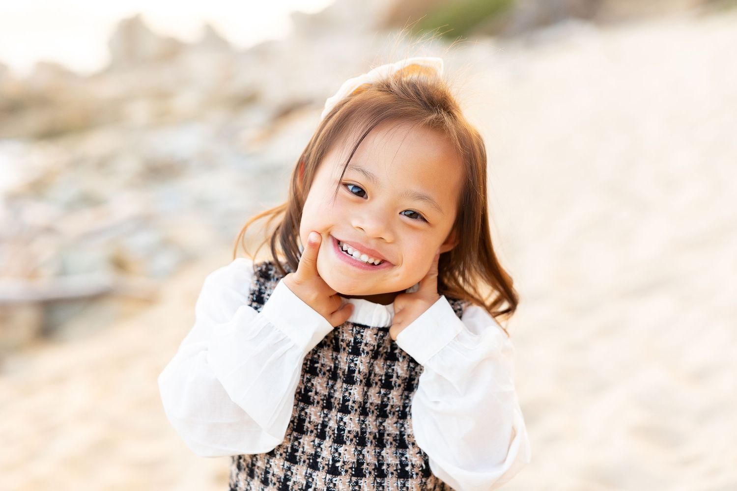 Child wearing a houndstooth dress and white sweater smiles joyfully while playing on the beach.