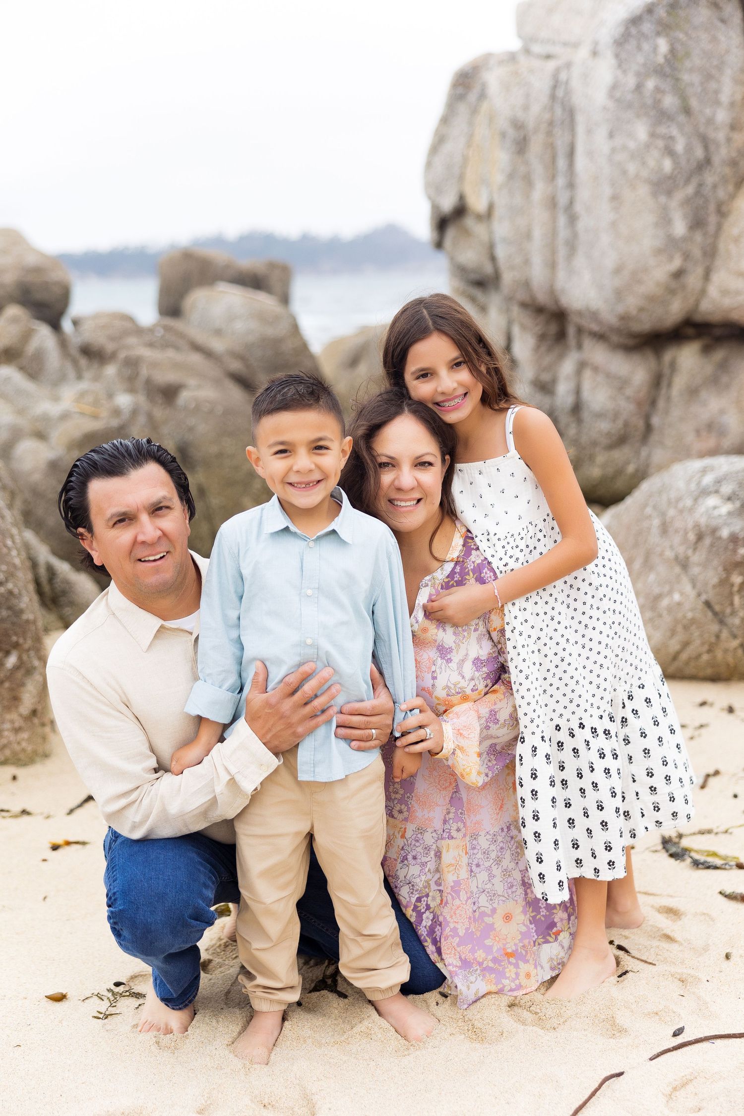 A happy family sits together on a sandy beach with large rocks in the background.