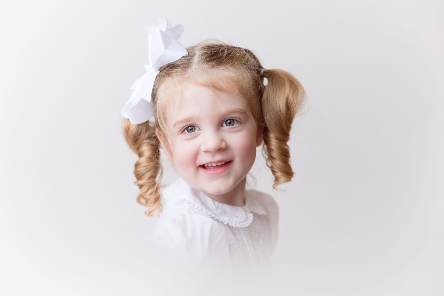 A studio portrait with white background showing curly pigtails and white bow hair accessories, heirloom portrait.