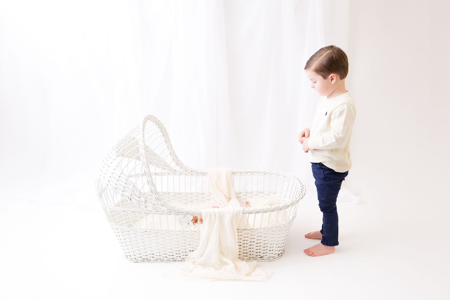 Small child in white sweater standing beside vintage white wicker bassinet.