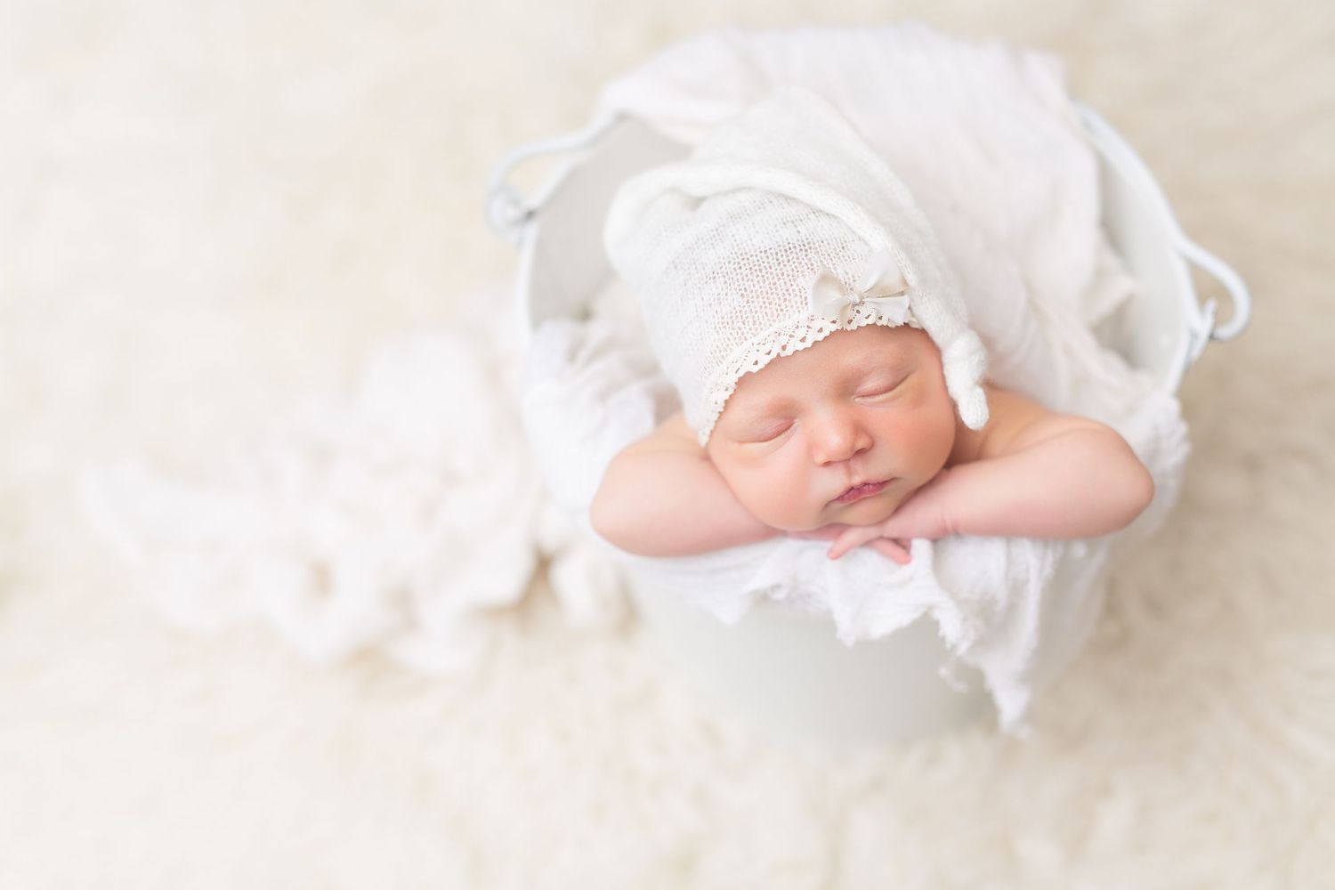 Sleeping newborn wrapped in white lace wearing delicate bonnet on white background.