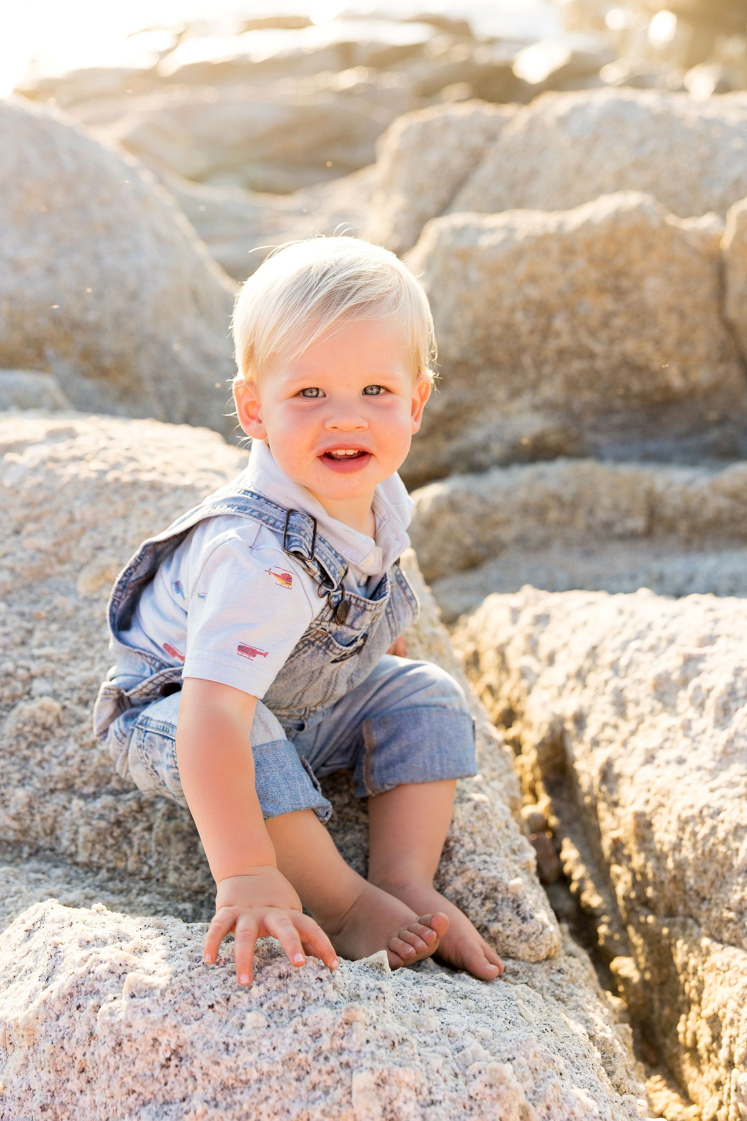 Baby in denim exploring rocky beach terrain during sunset photoshoot.