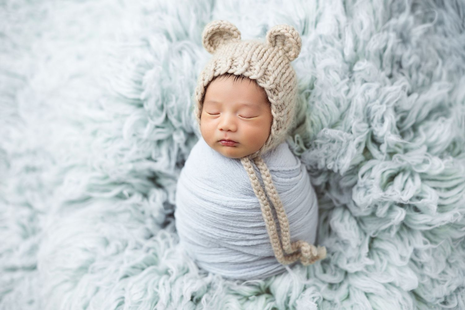 Sleeping newborn wrapped in gray swaddle wearing knit bear hat on blue fluffy rug.