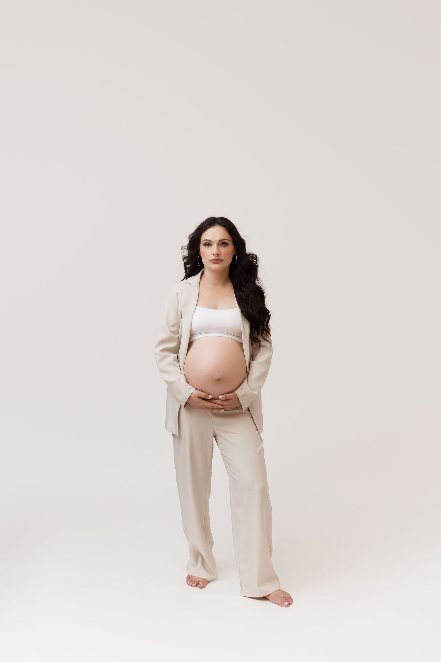 Studio maternity photo of woman in open blazer and bra gently holding her belly on light background in Pacific Grove