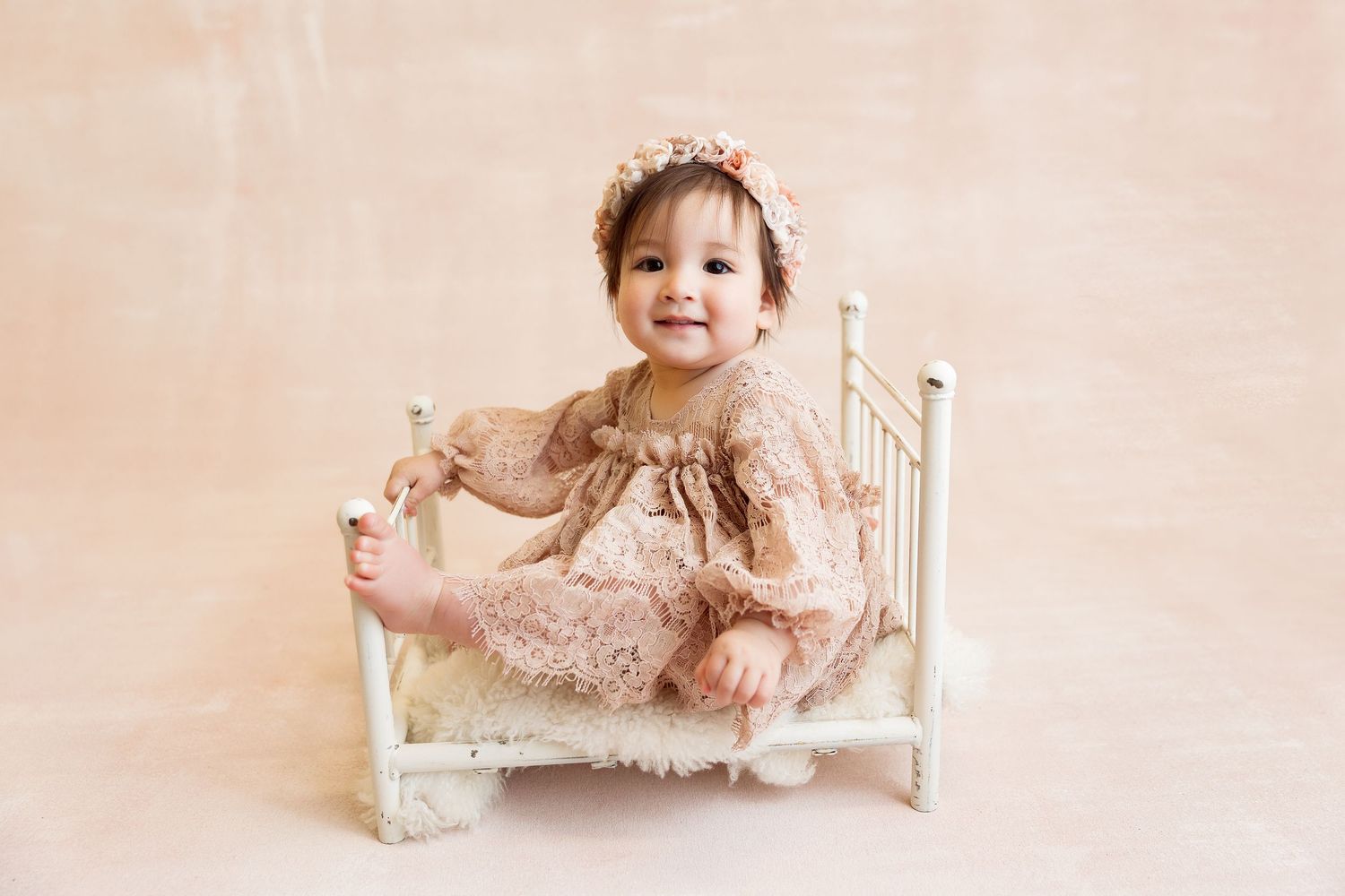 Baby wearing peach dress sits in miniature white bed during photo shoot.