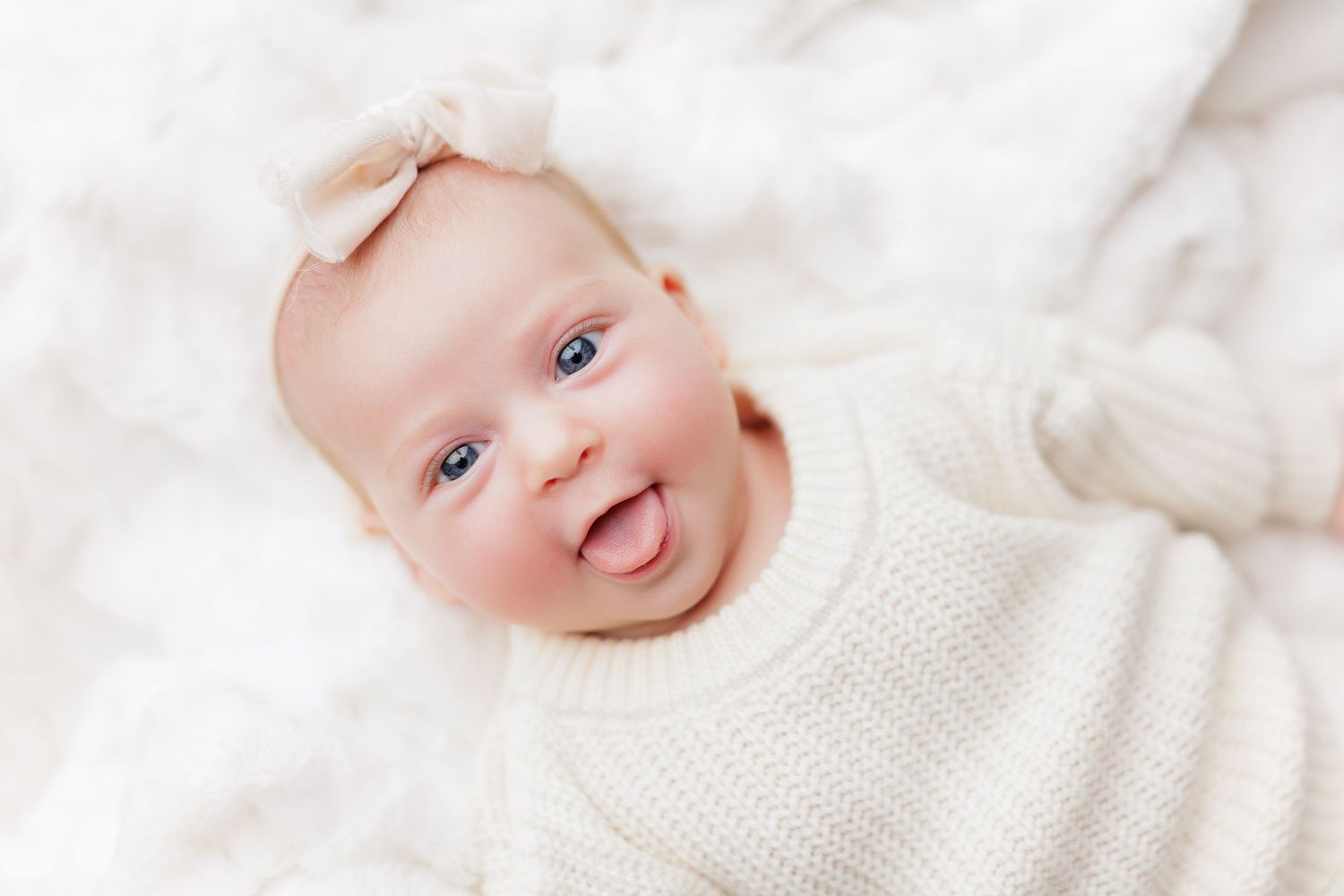 A baby wearing a white knit sweater smiles while lying on soft white bedding.