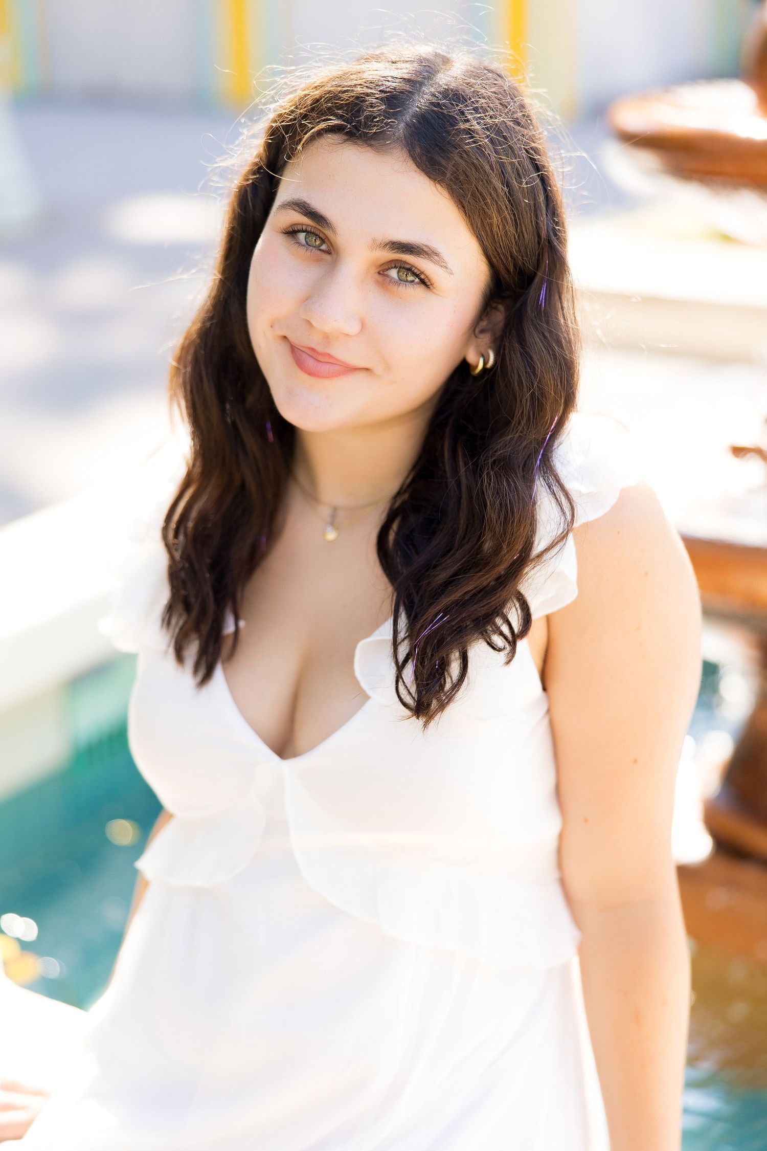 Young woman with dark hair in white dress smiling near a pool fountain in bright sunlight.
